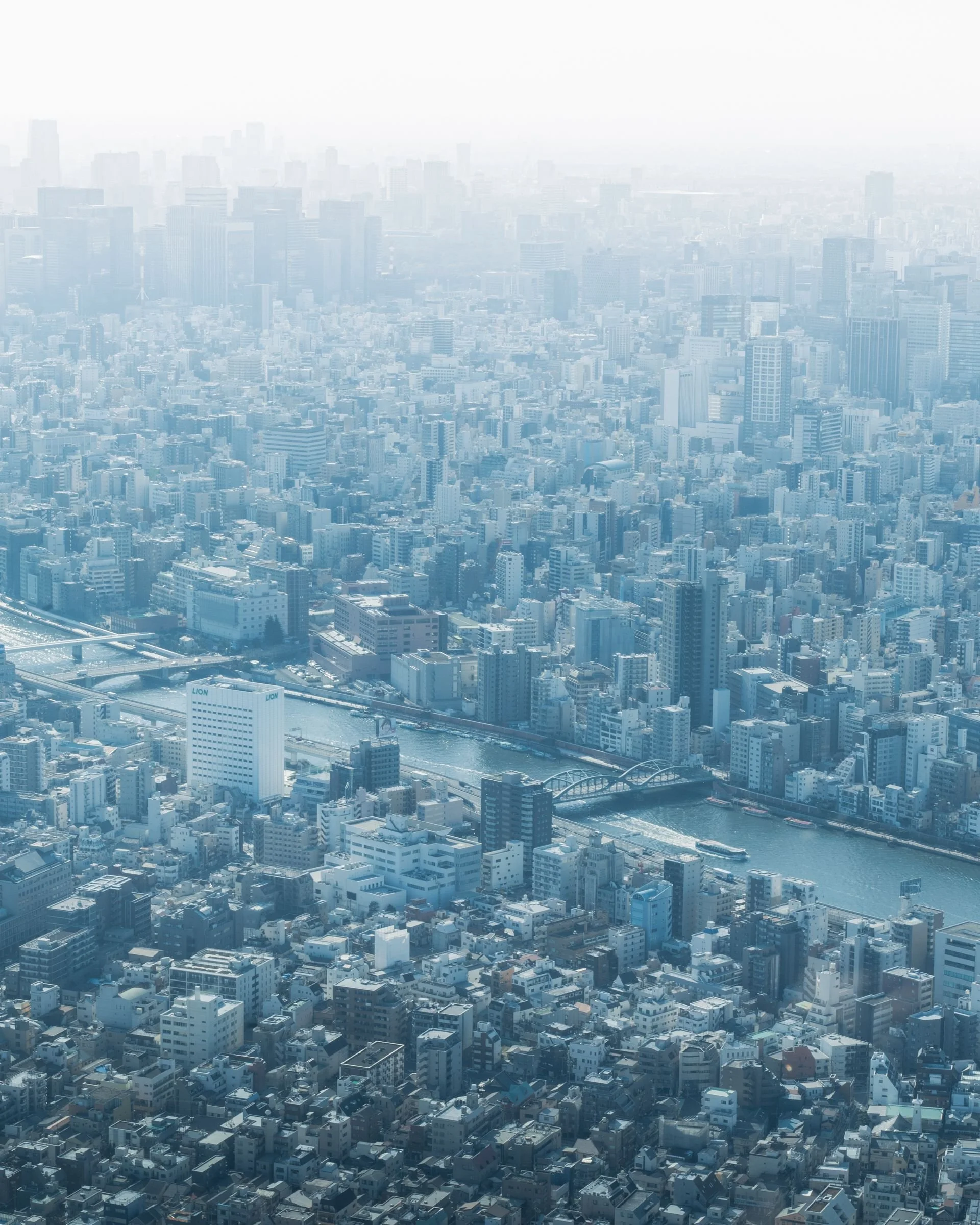 Aerial view of a dense cityscape with high-rise buildings, a river running through the city, and bridges, under a hazy sky.