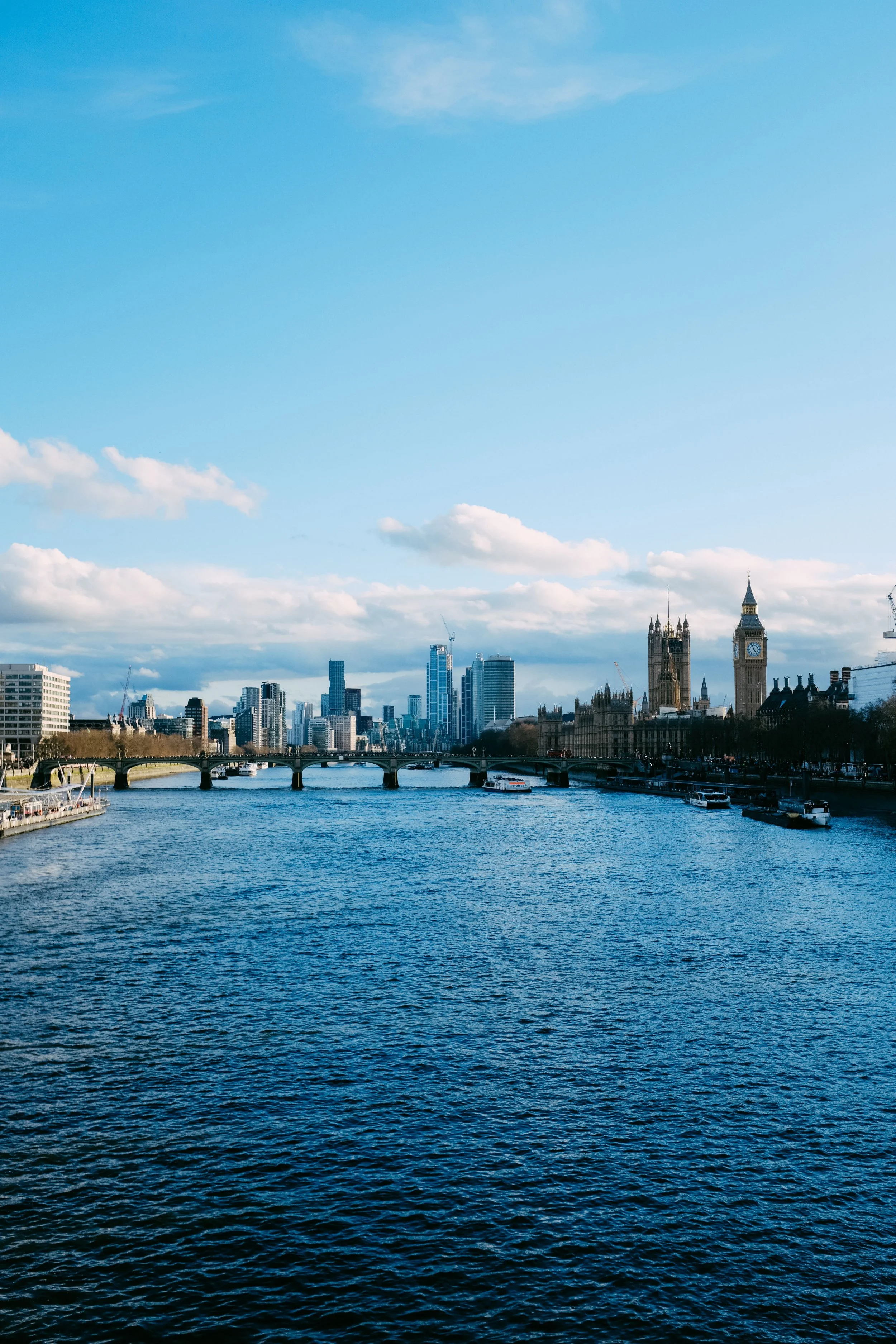 City skyline with tall buildings and the Big Ben clock tower in London, seen across a river with boats and a bridge, under a partly cloudy sky.
