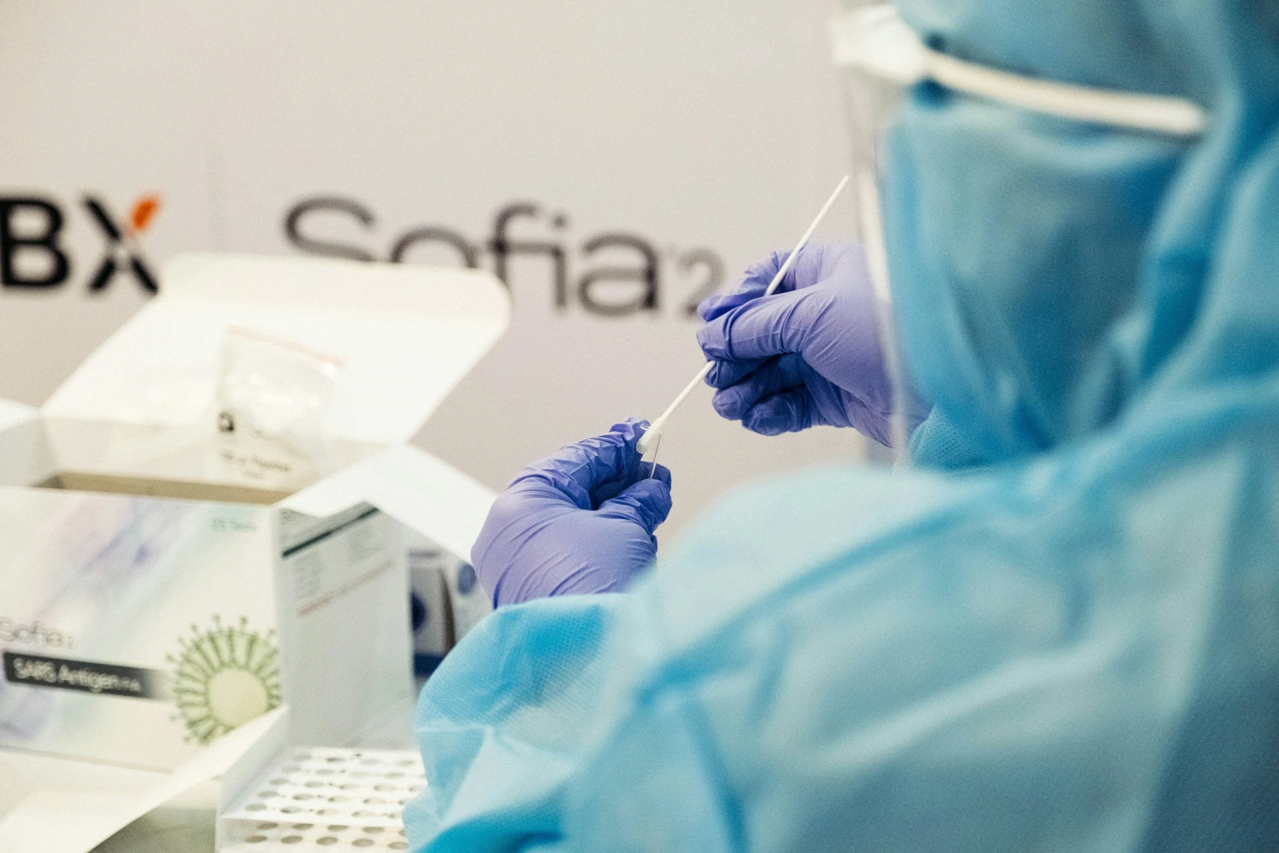 A healthcare worker in blue protective gear and purple gloves is handling a cotton swab for a medical test, with a box of testing supplies in the background. Gut health testing.