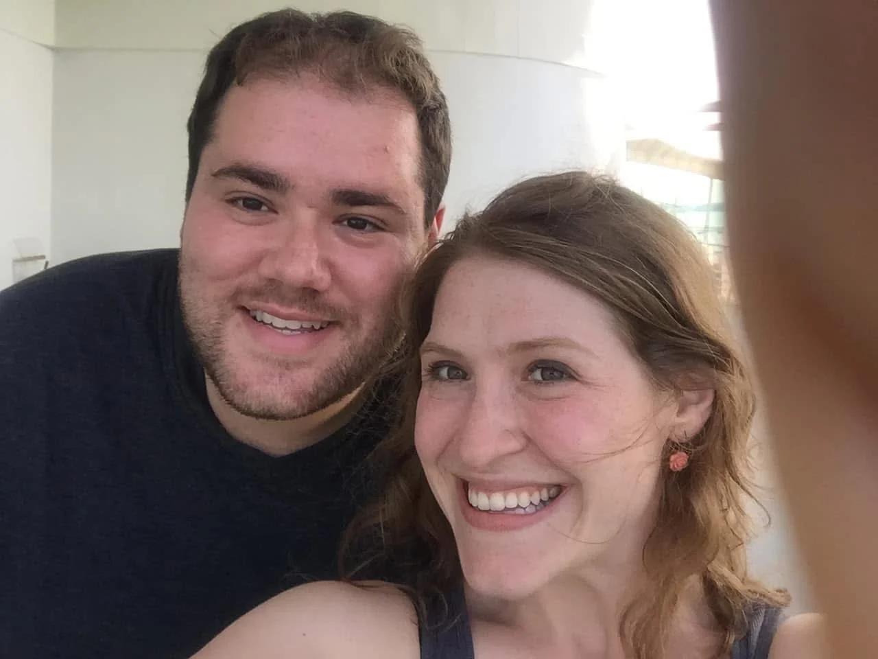 A smiling man and woman taking a selfie together indoors, with natural light coming from windows behind them.
