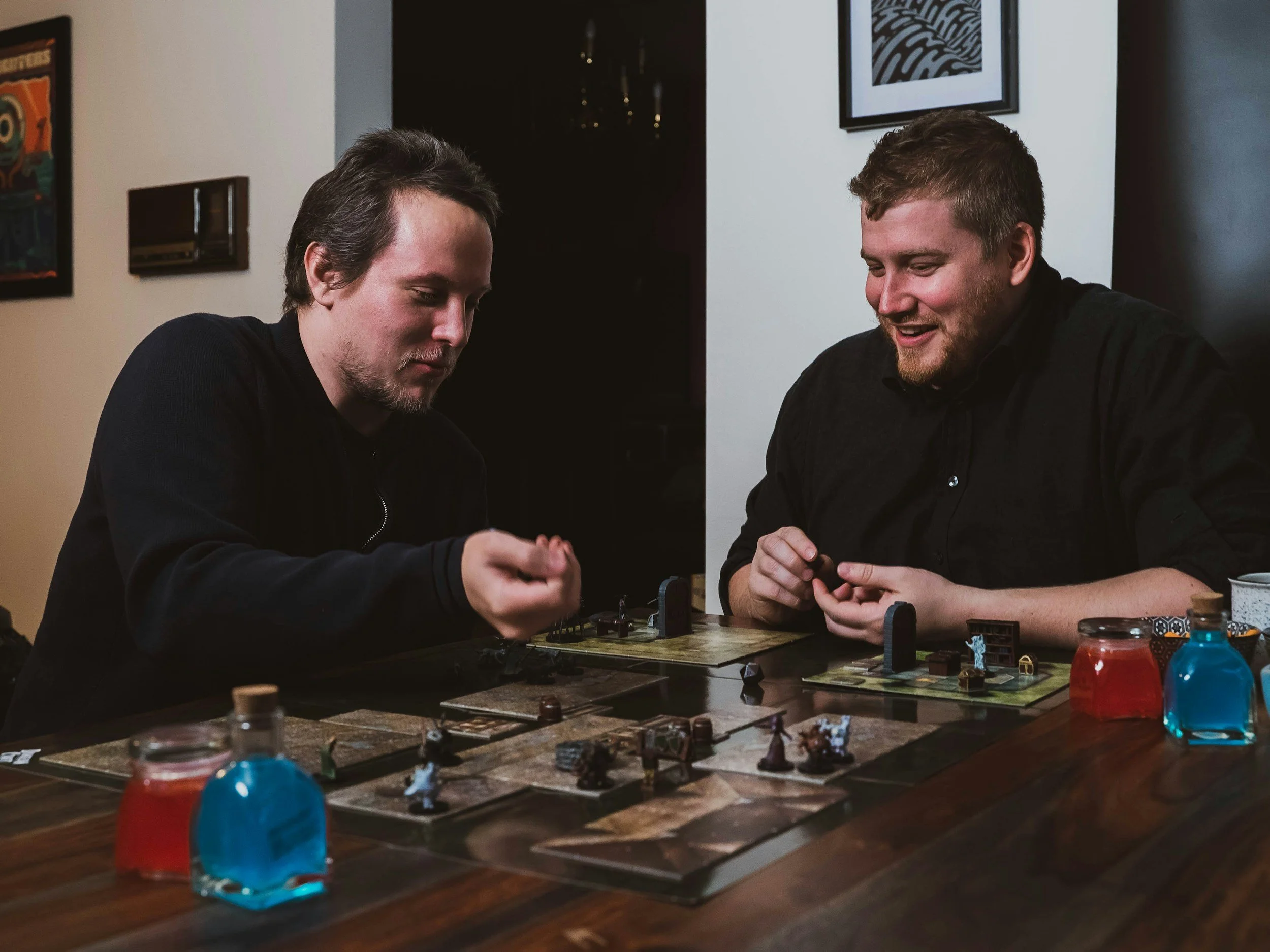 Two men playing a tabletop strategy game with miniatures, surrounded by game pieces and color-coded fire bottles, in a room with artwork on the walls.