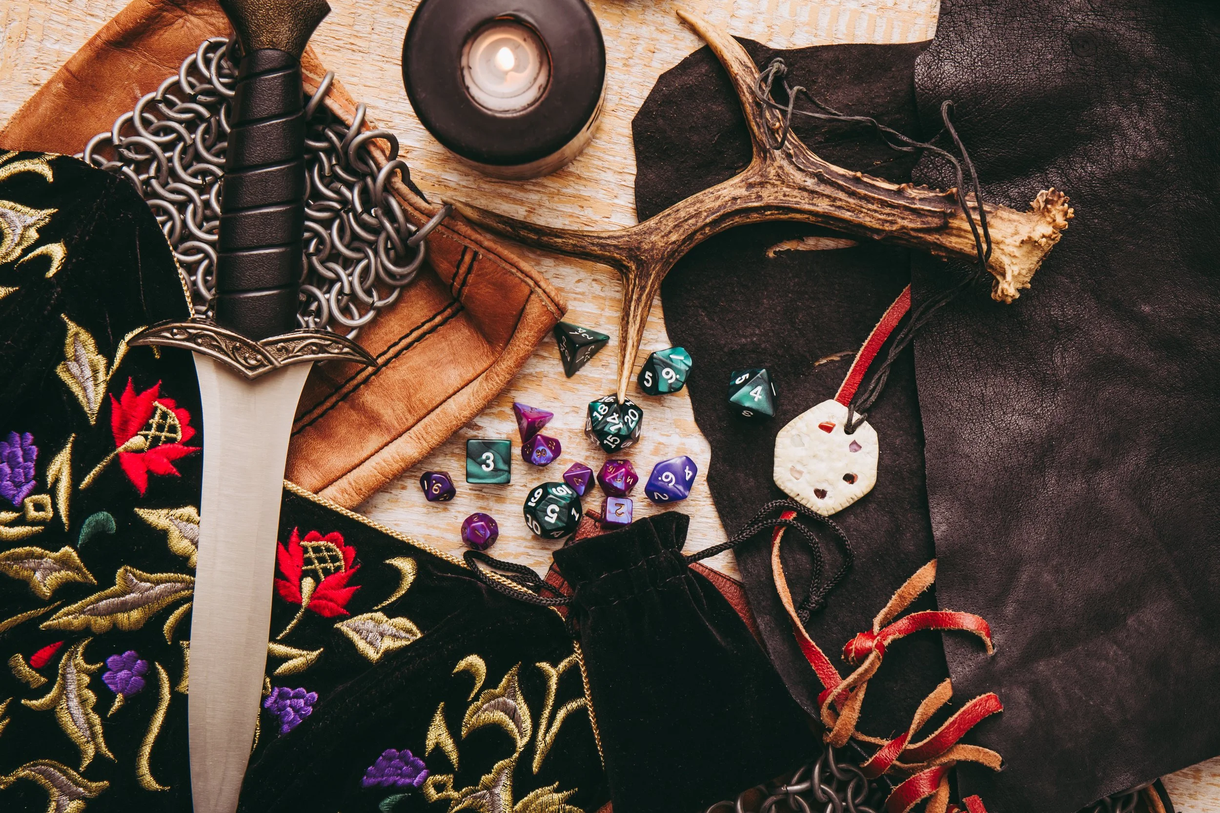 A collection of fantasy role-playing game items on a wooden surface, including a dagger, a candle, a deer skull, fabric, a moonstone pendant, and various colorful polyhedral dice.