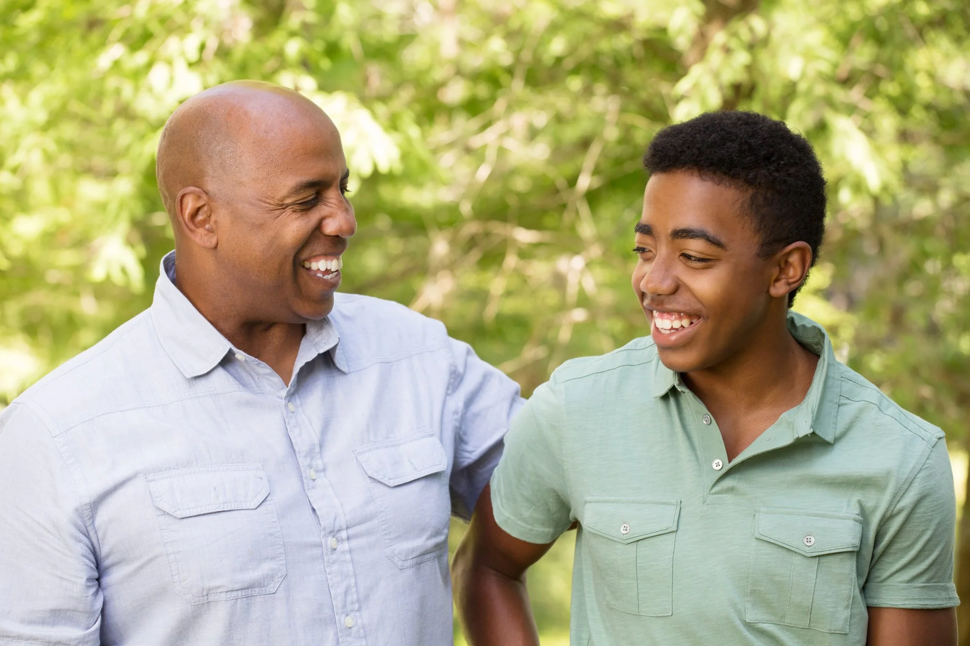 Un hombre de mediana edad y un adolescente, sonriendo y charlando al aire libre entre árboles frondosos.