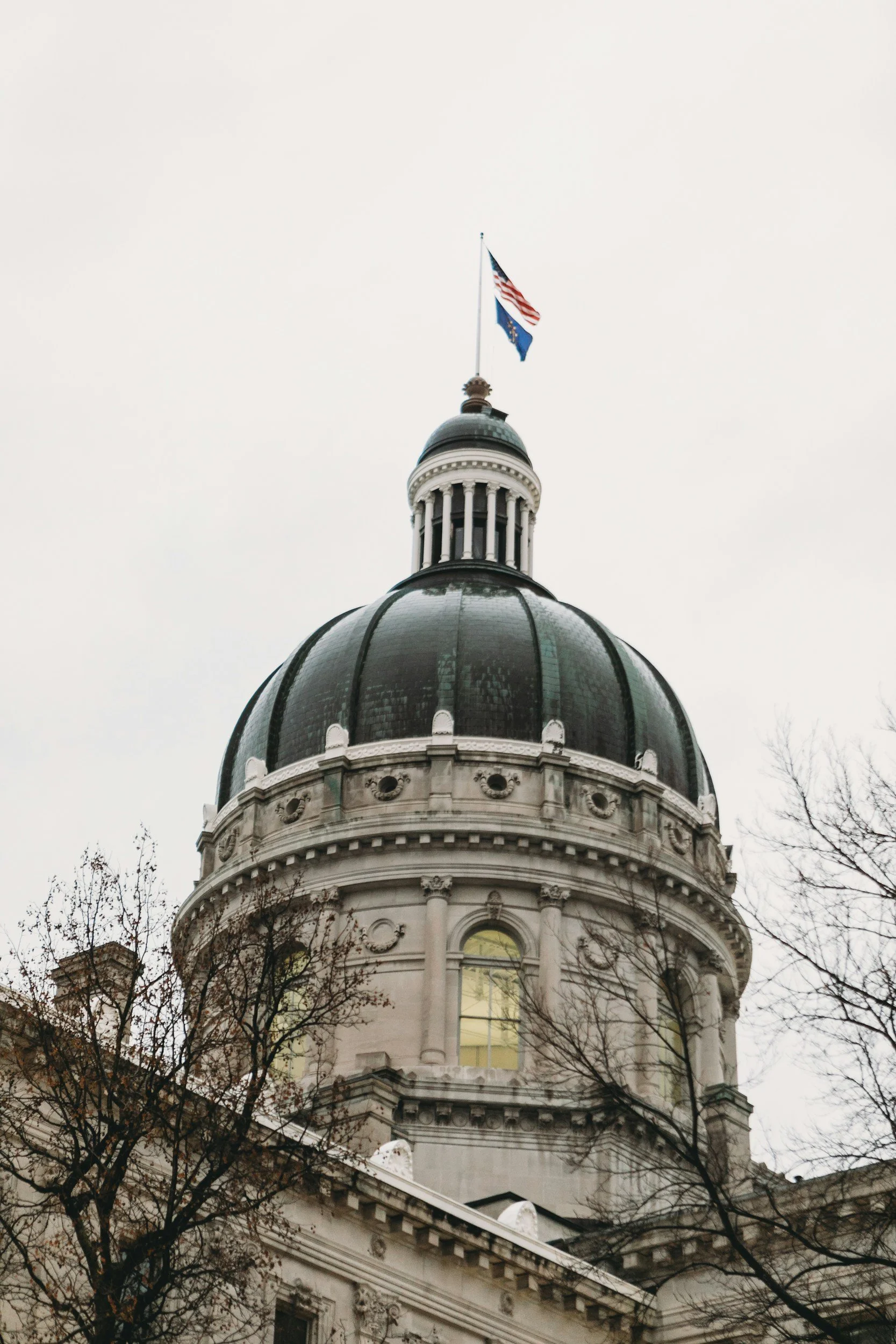 Vista del edificio del Capitolio del estado de Indiana, con una gran cúpula y un mástil en lo alto en el que ondean la bandera estadounidense y la bandera estatal, con un cielo nublado de fondo y árboles sin hojas en primer plano.
