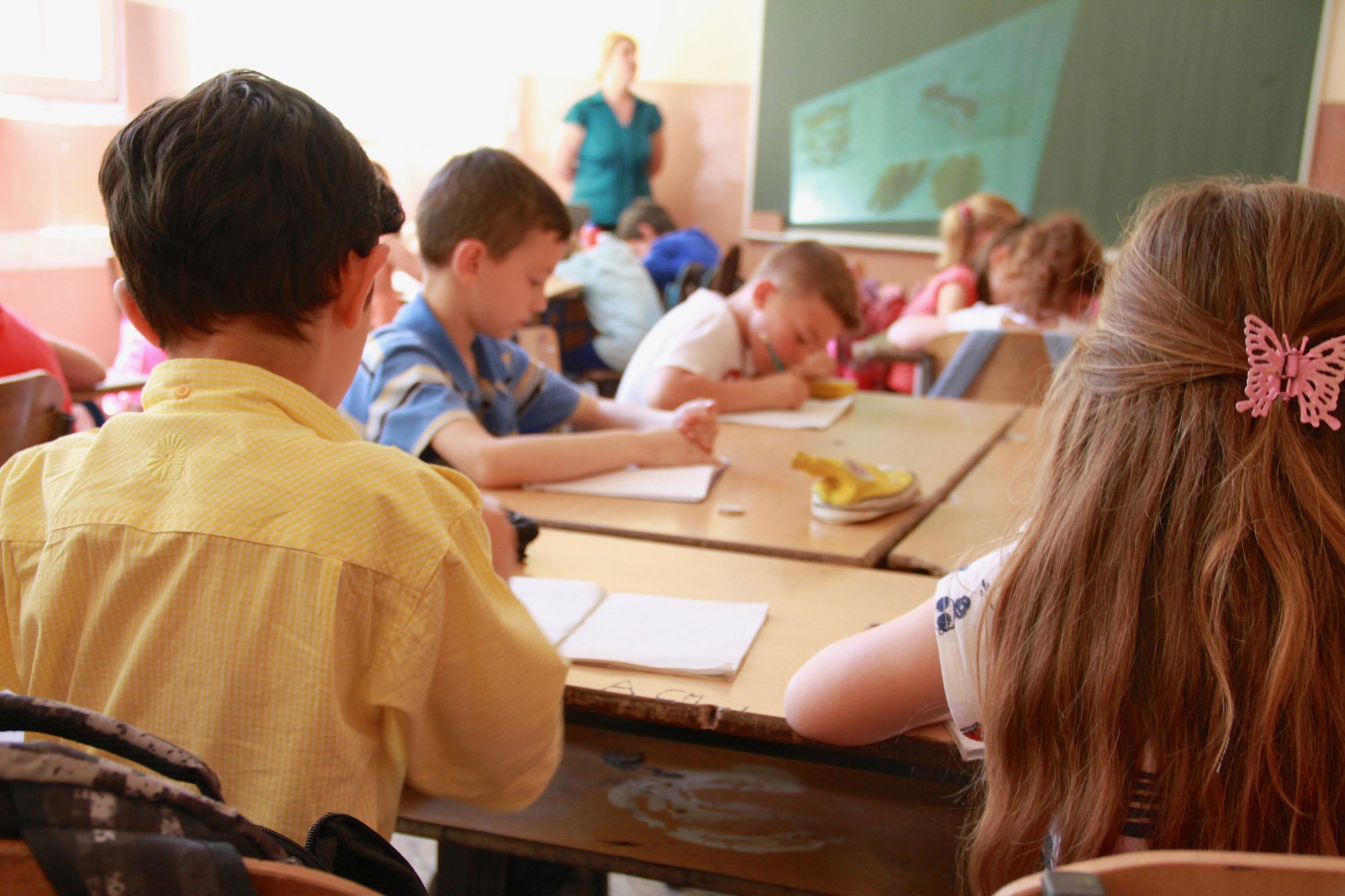 Children sitting at desks writing, teacher standing at front of classroom, blackboard on wall.