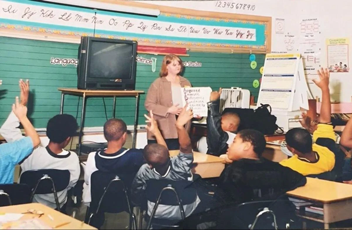 Un aula de la década de los 90 en la que un profesor está de pie frente a los alumnos, que tienen la mano levantada, y sostiene una cartulina. Varios alumnos tienen la mano levantada, sentados en sus pupitres, en un aula con pizarra verde y carteles educativos.