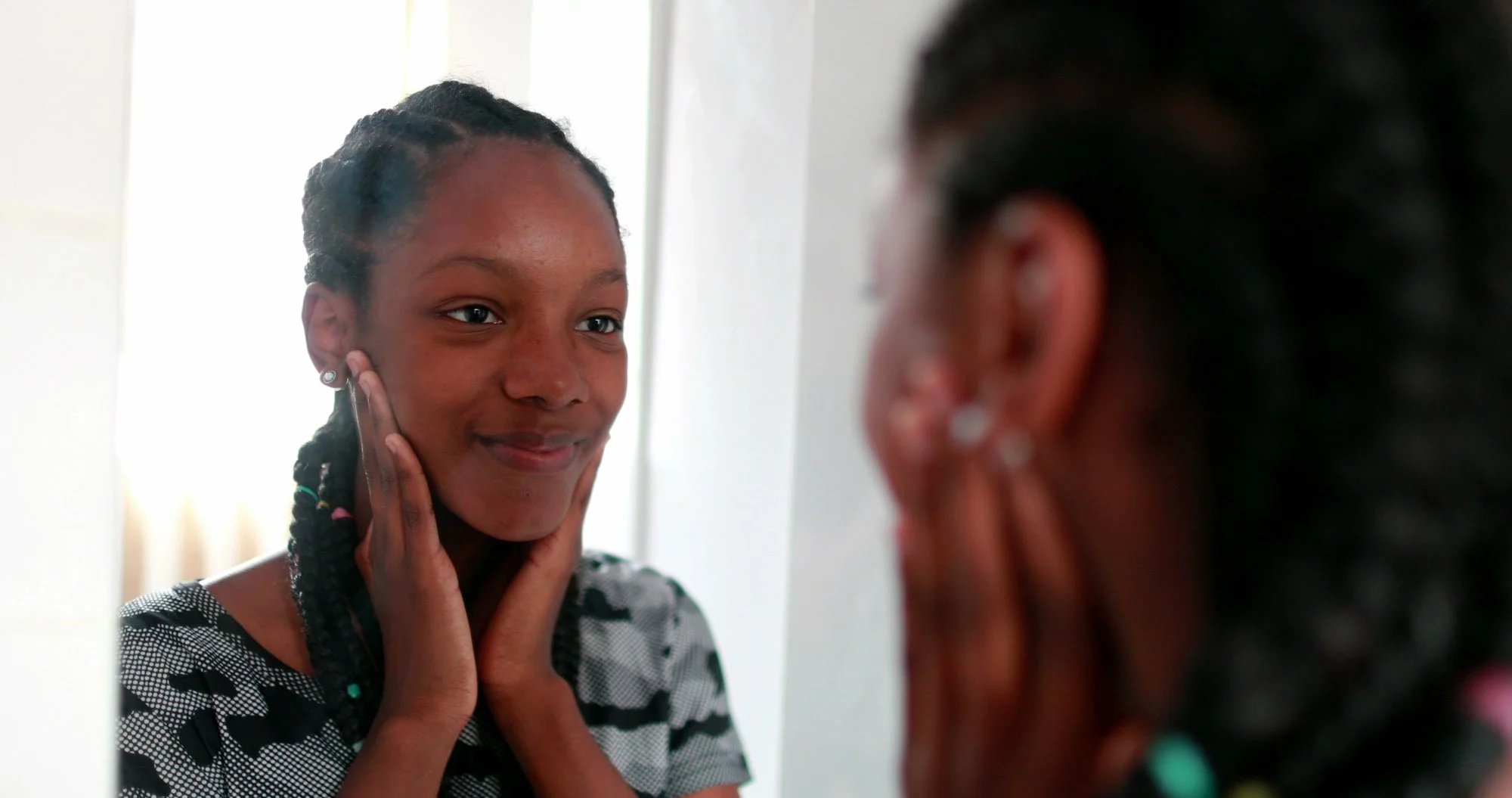 Una joven adolescente se mira en el espejo, acariciándose suavemente la cara y sonriendo con dulzura, con una camiseta estampada en blanco y negro y unos pendientes.