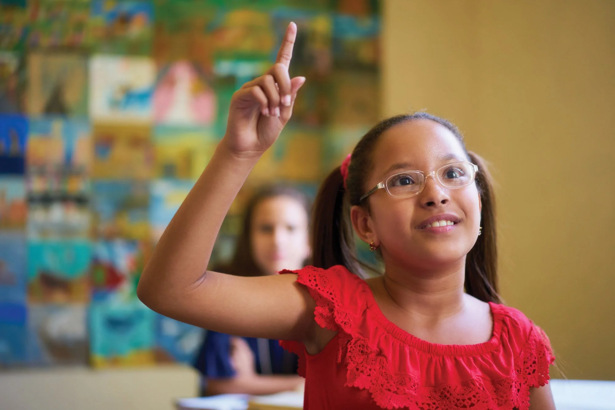 Una niña con gafas y un vestido rojo está levantando la mano derecha con el dedo índice apuntando hacia arriba. Está sonriendo y sentada delante de un fondo colorido.