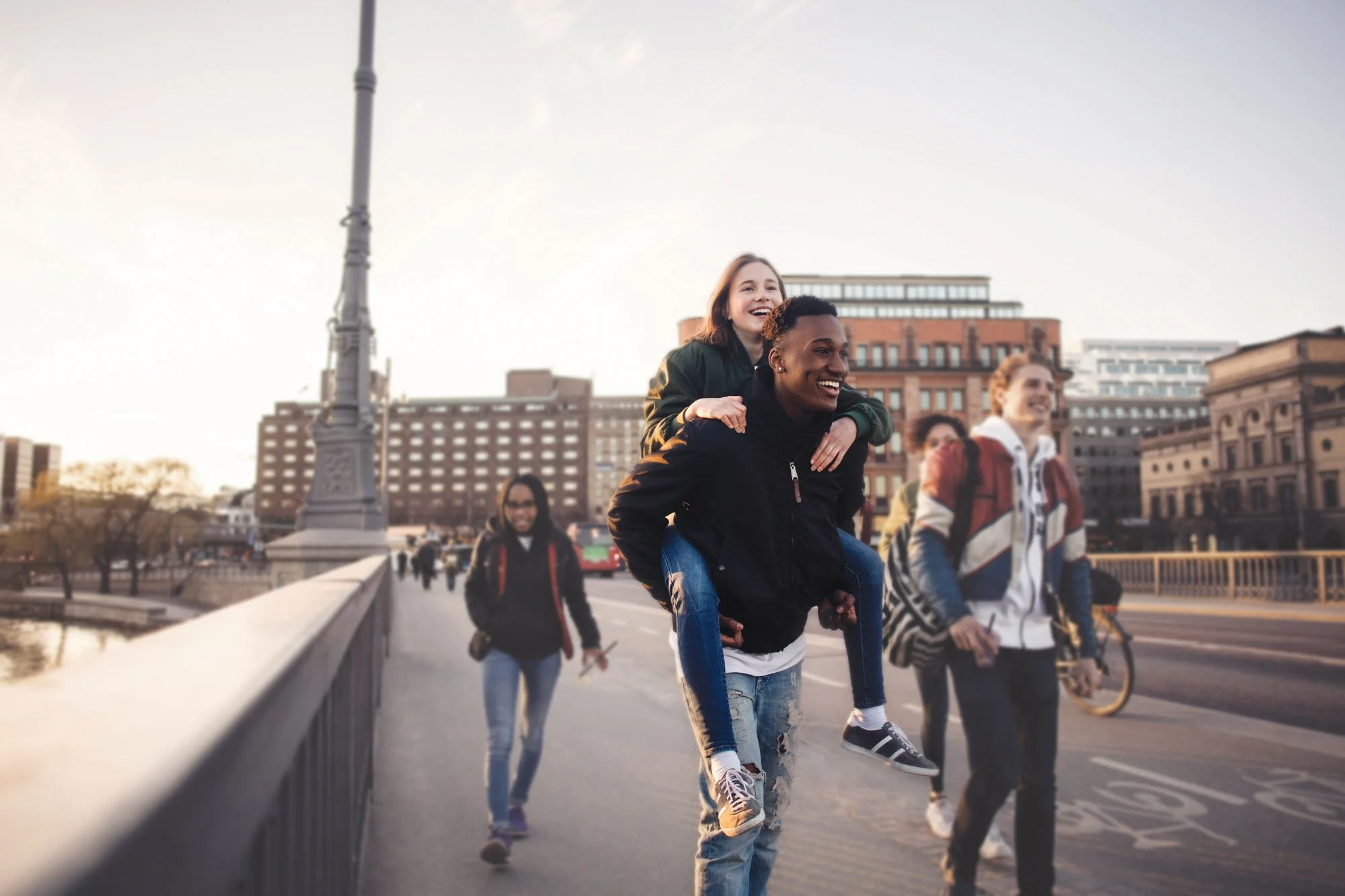 Un grupo de jóvenes de diferentes orígenes que pasean y se divierten montados a caballito en un puente de la ciudad al atardecer, con edificios y una farola al fondo