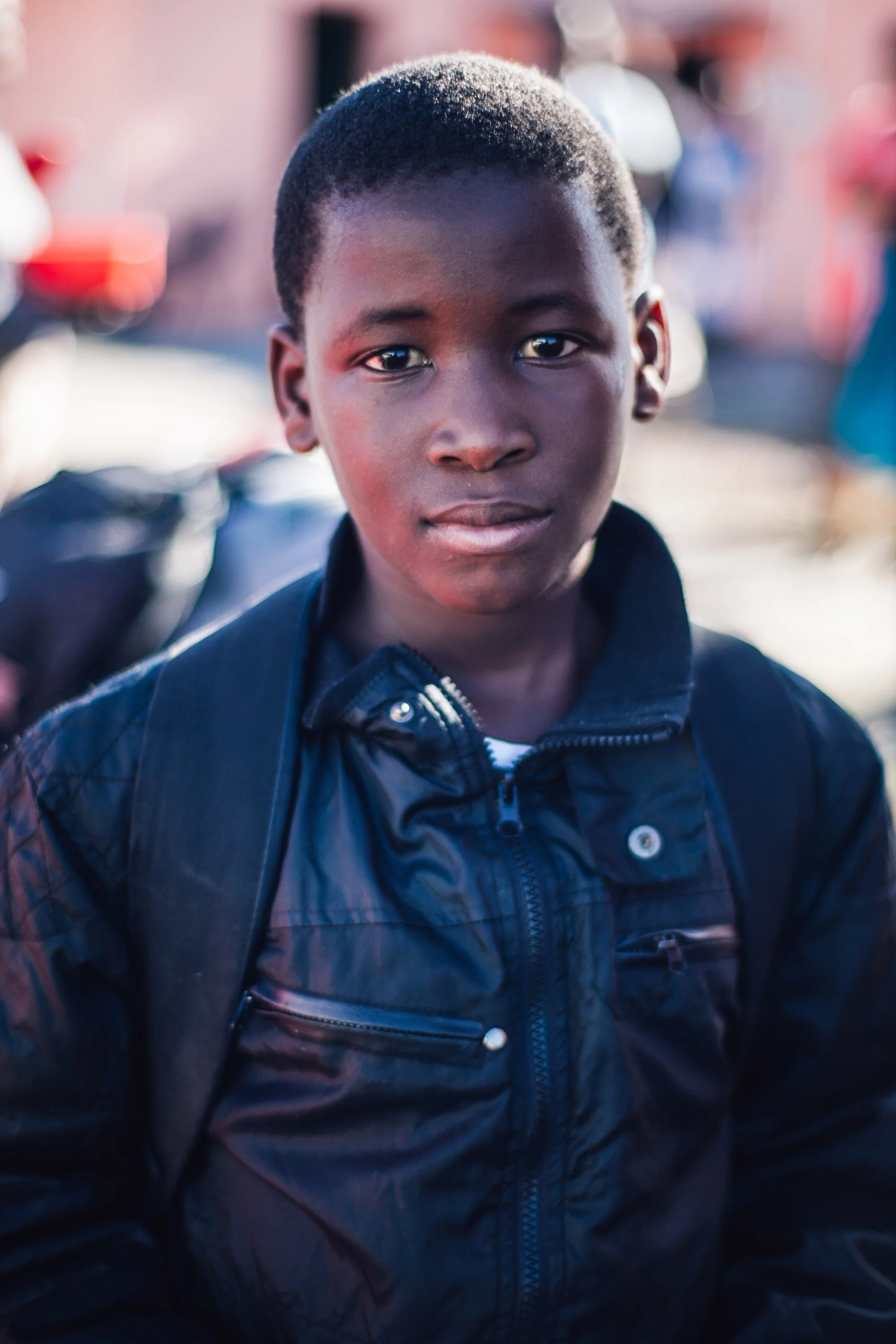 Primer plano de un niño negro con el pelo corto, vestido con una chaqueta negra, de pie al aire libre con un fondo desenfocado.