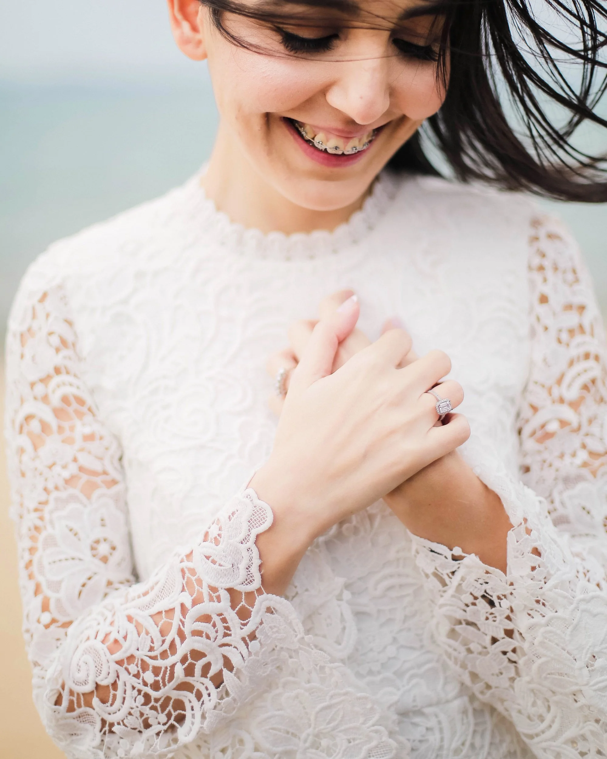 Close-up of a smiling teenage girl with dark hair and braces, wearing a white lace dress, holding her hands near her chest.