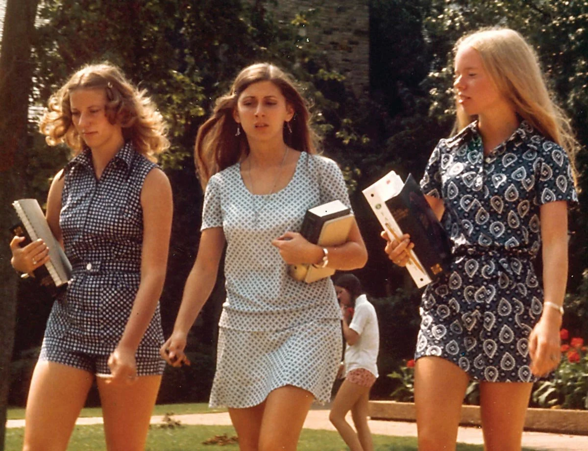 Fotografía vintage de la década de los 70 en la que aparecen tres adolescentes caminando al aire libre, cada una con cuadernos y papeles en la mano; dos de ellas tienen el pelo rubio y una, castaño, y llevan vestidos cortos estampados.