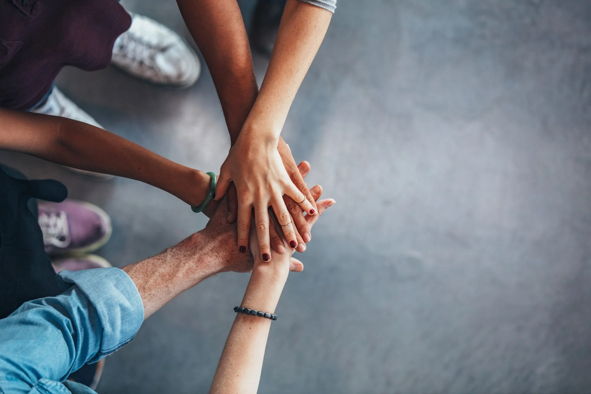 Un grupo de personas de diversa procedencia con las manos unidas en el centro, en señal de unidad y trabajo en equipo, sobre un suelo gris.