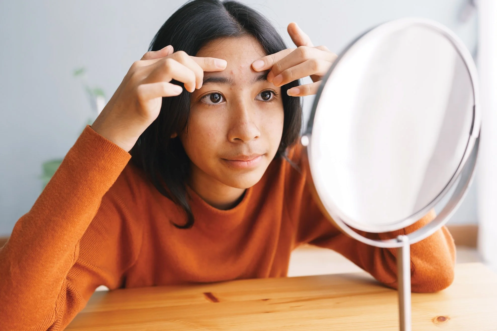Una adolescente o preadolescente de pelo oscuro se mira la frente en un espejo, examinándose la piel. Lleva un jersey naranja y está sentada en una mesa de madera.