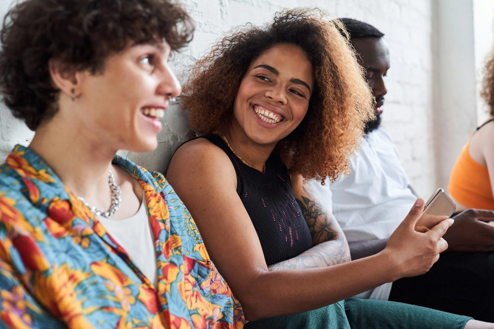 Youth sitting in a row, smiling and laughing together, some holding phones, against a white brick wall.