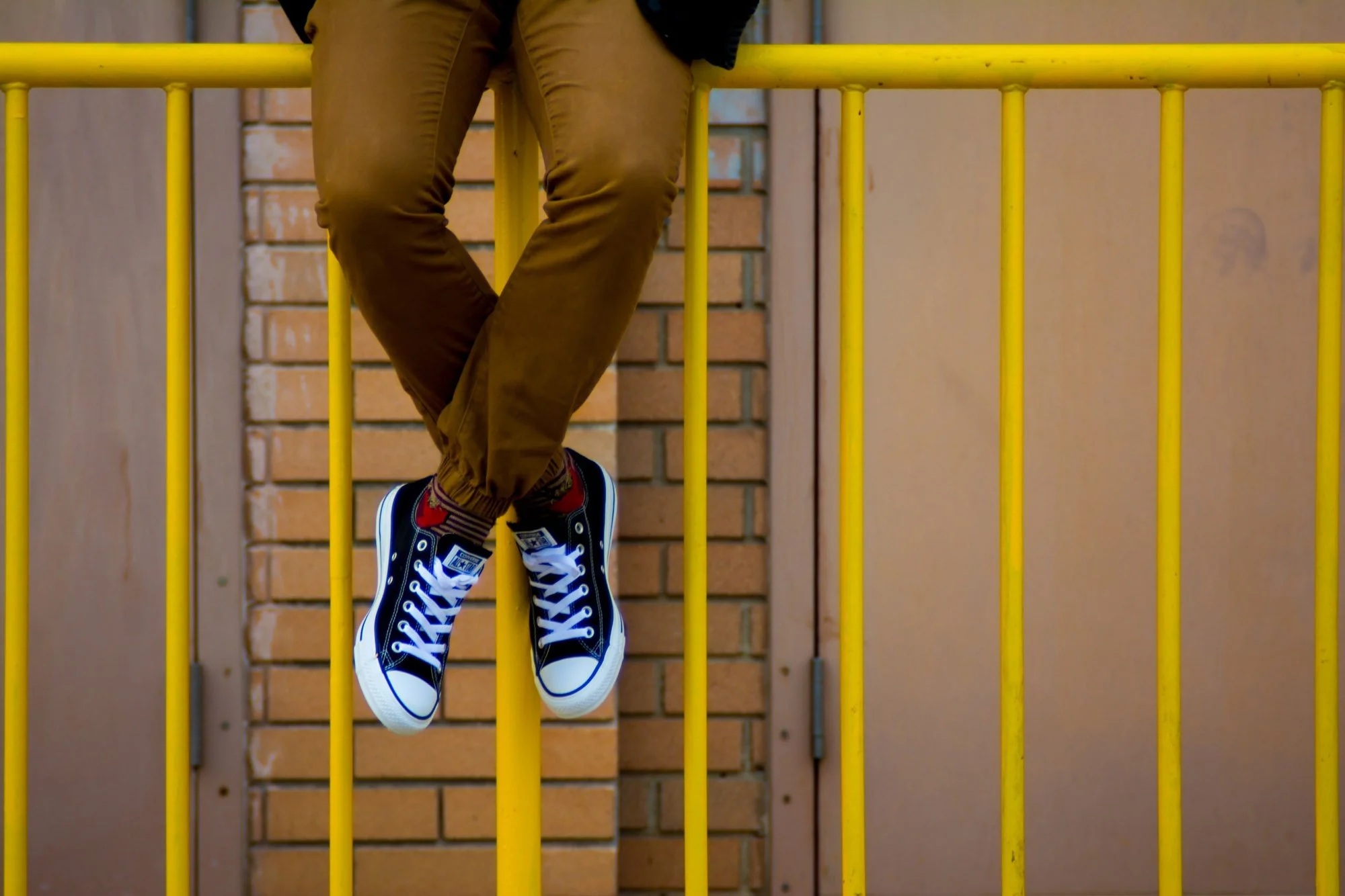 Teen wearing tan pants and black jacket sitting on yellow metal railing with legs crossed, wearing blue and white Chuck Taylors, against a background of brick wall and beige surface.