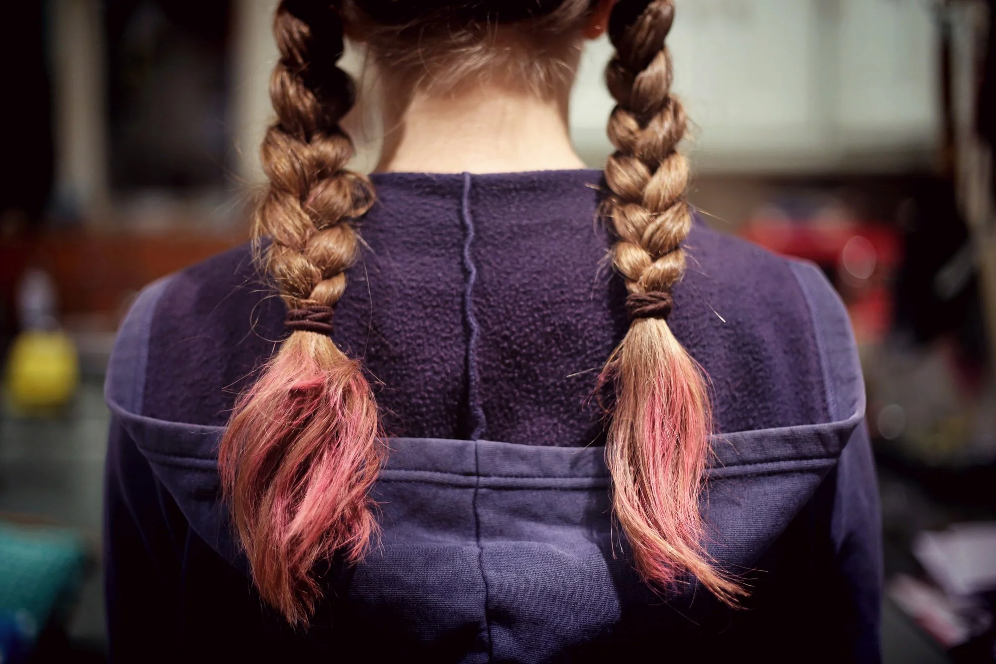 La espalda de una niña con dos trenzas largas con las puntas teñidas de rosa, que lleva una chaqueta morada, con un fondo borroso.