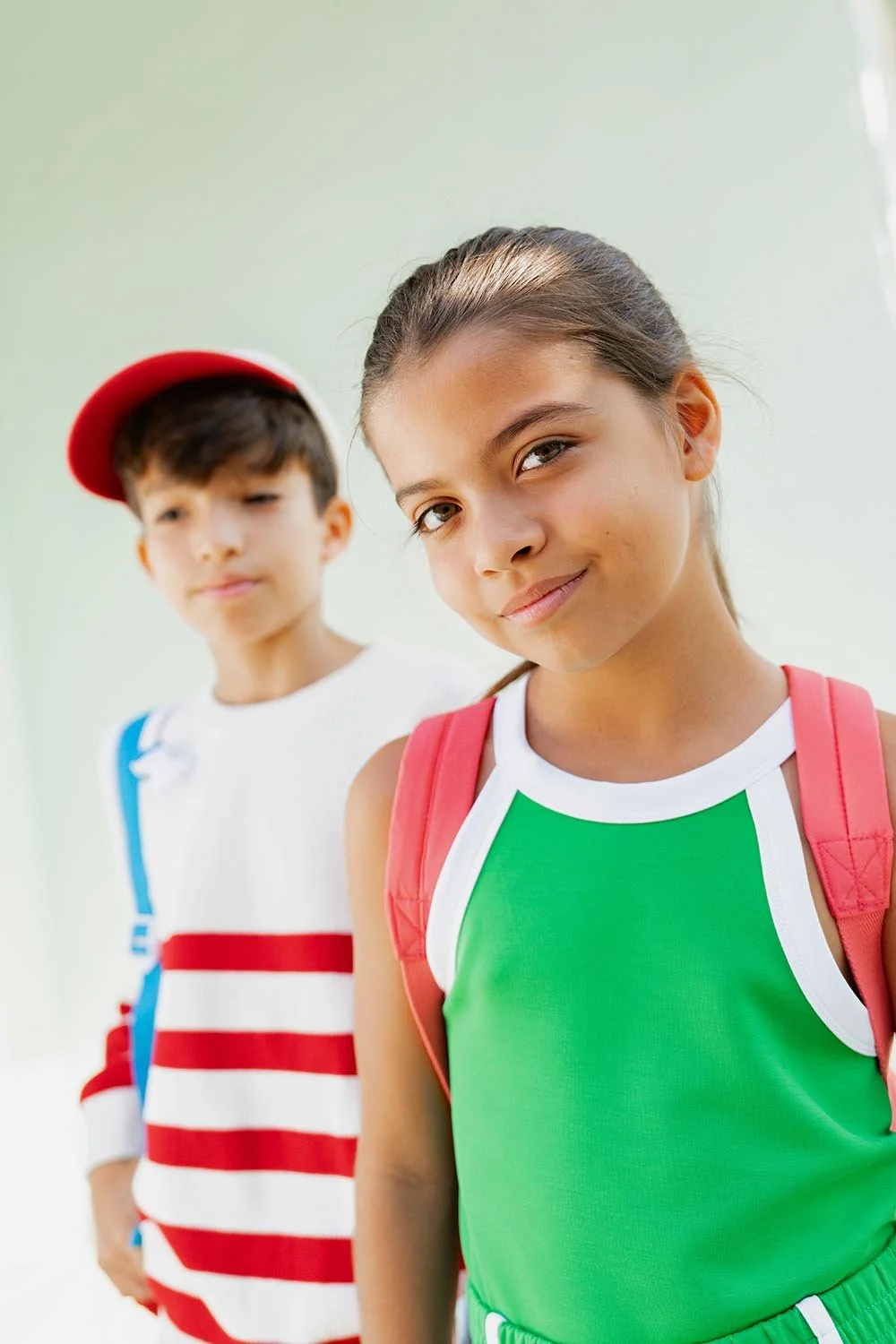 Una niña con una camiseta verde y una mochila que sonríe a la cámara, y un niño con una mochila de pie detrás de ella; ambos llevan ropa informal de verano.