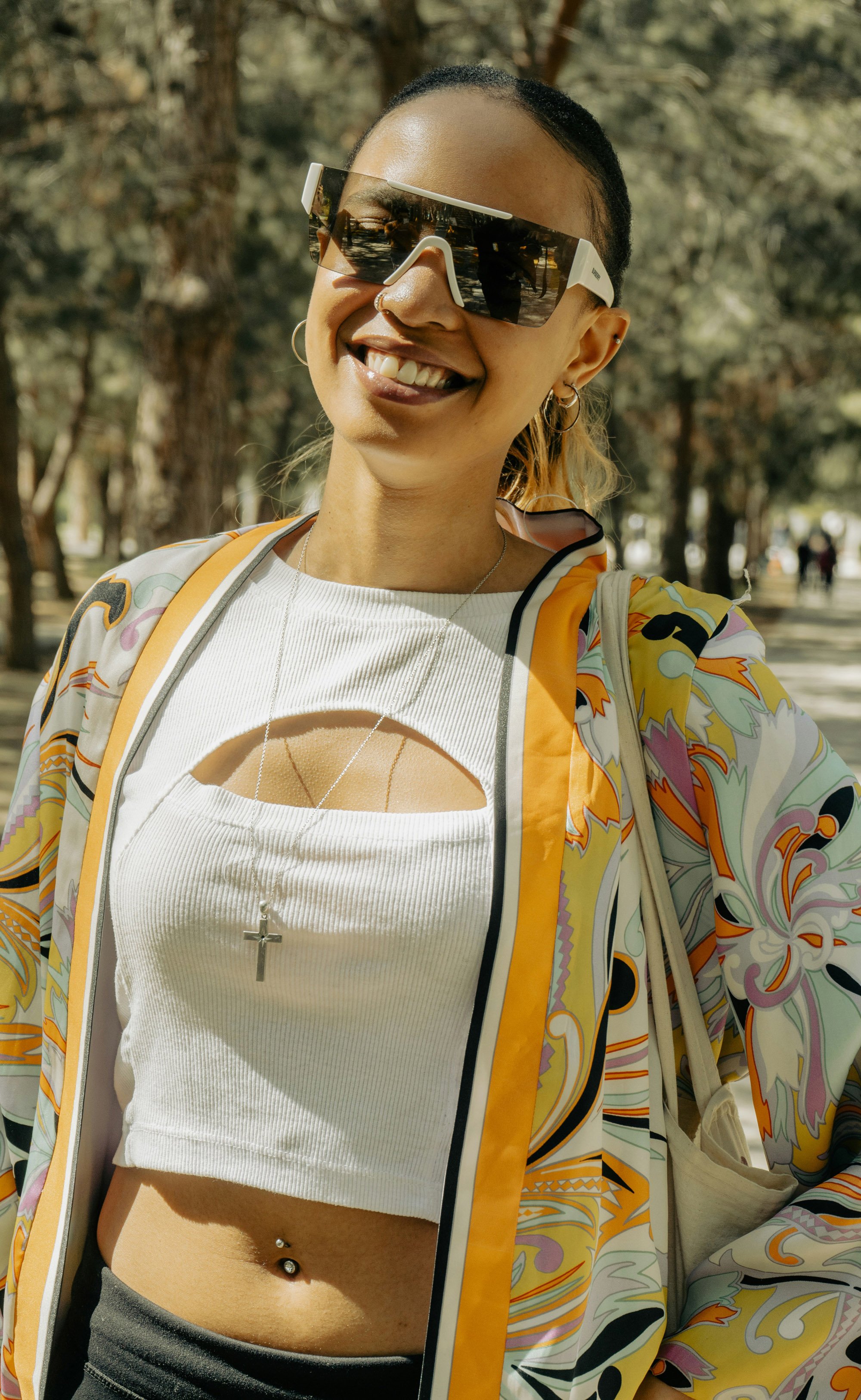 Mujer sonriente con gafas de sol de marca, de pie al aire libre en un parque con árboles al fondo.