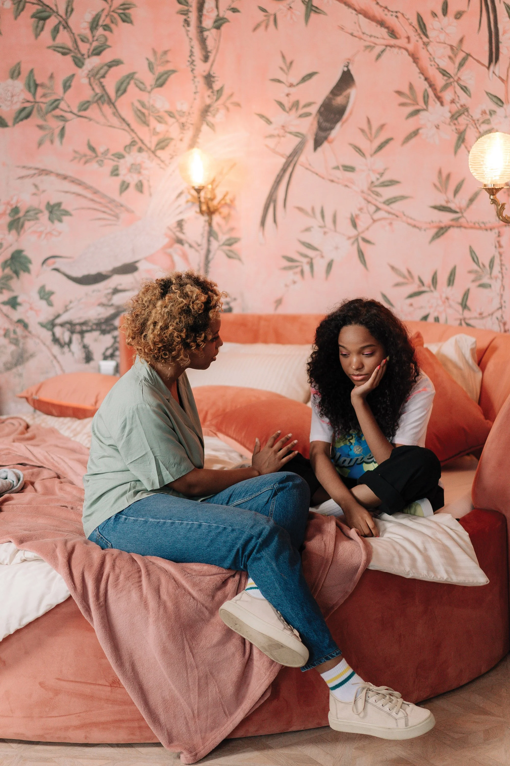 Una madre y su hija adolescente están sentadas en una cama y charlan. La habitación tiene una iluminación cálida, ropa de cama rosa y una pared con un diseño botánico en el que aparecen pájaros y flores.