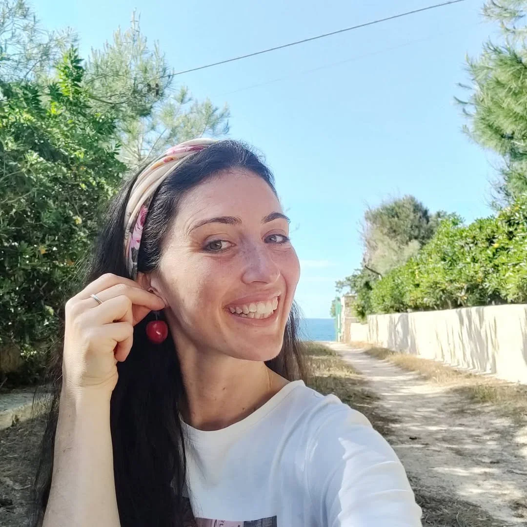 A woman with dark hair, wearing a headscarf, smiling and holding a cherry earring, standing on a dirt path with trees and bushes on either side, with a view of the sea in the background under a clear blue sky.