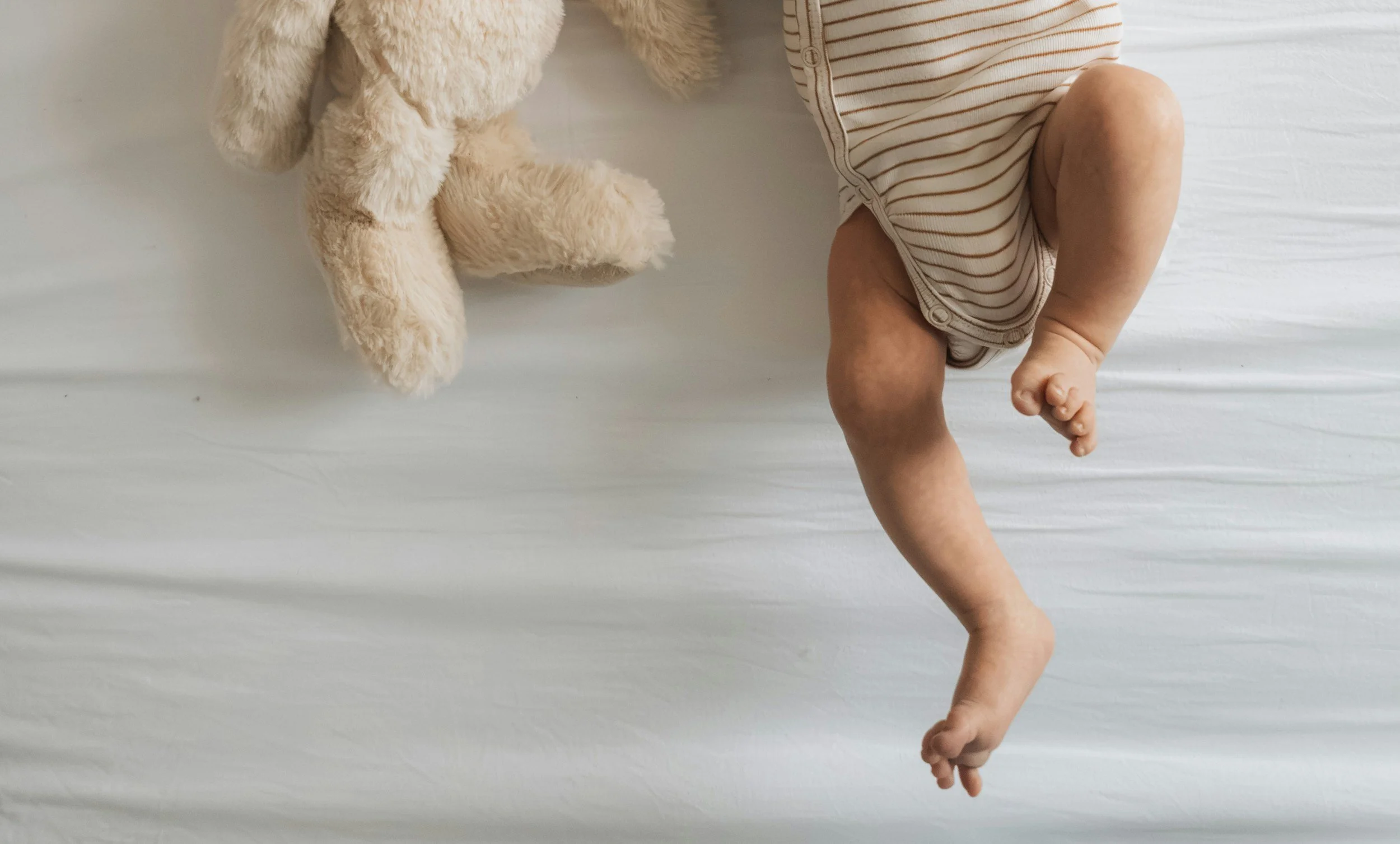 Newborn baby sleeping on a white surface, wearing a striped outfit, with a beige plush teddy bear nearby.