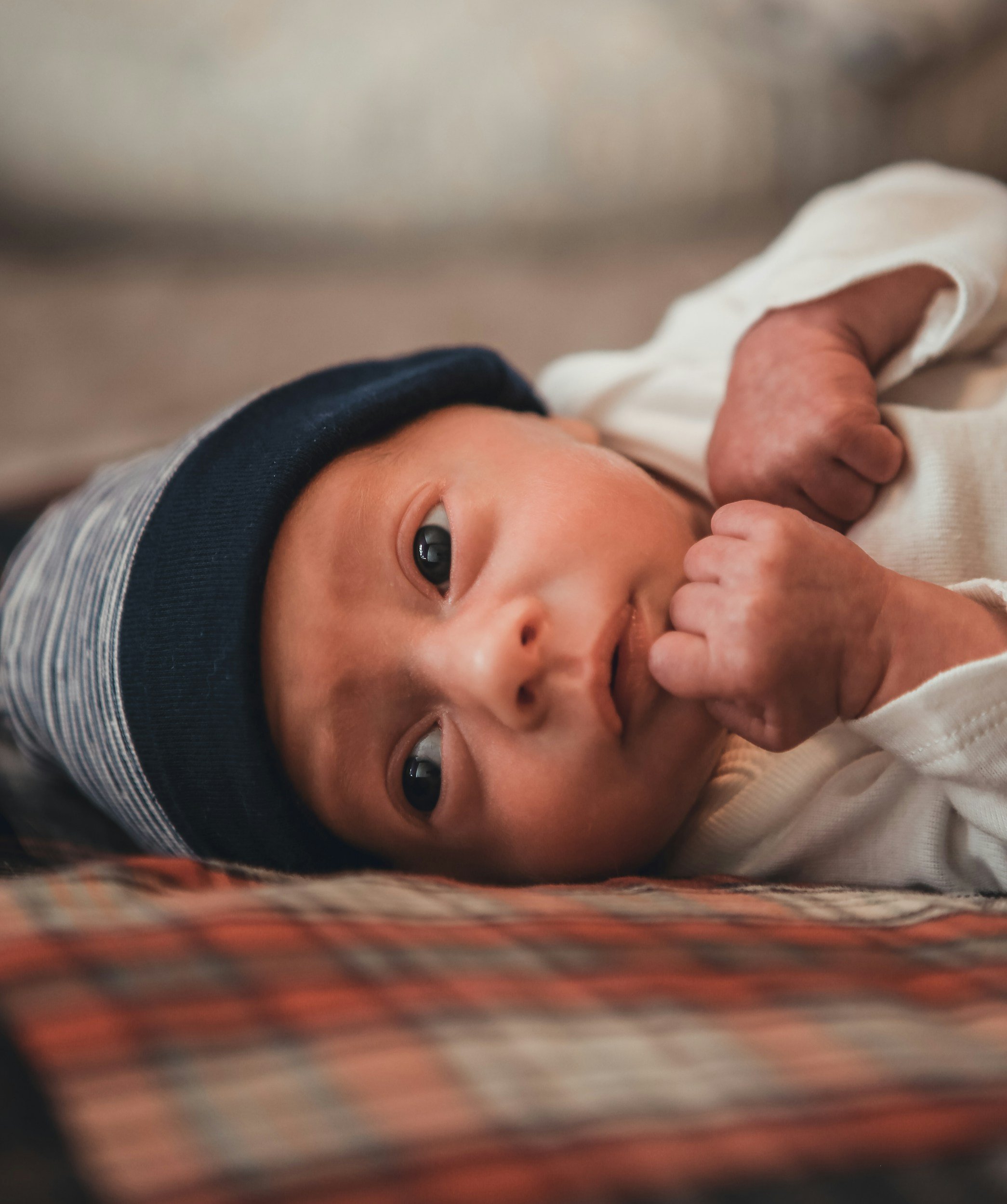 Close-up of a newborn baby lying on a blanket, wearing a striped hat and white outfit, looking at the camera with a hand near the face.