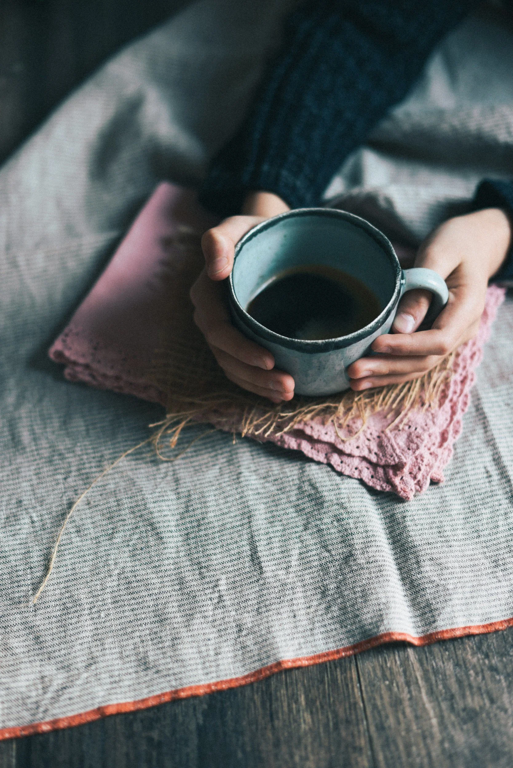 Person holding a ceramic mug of black coffee, sitting on a textured cloth on a wooden surface.