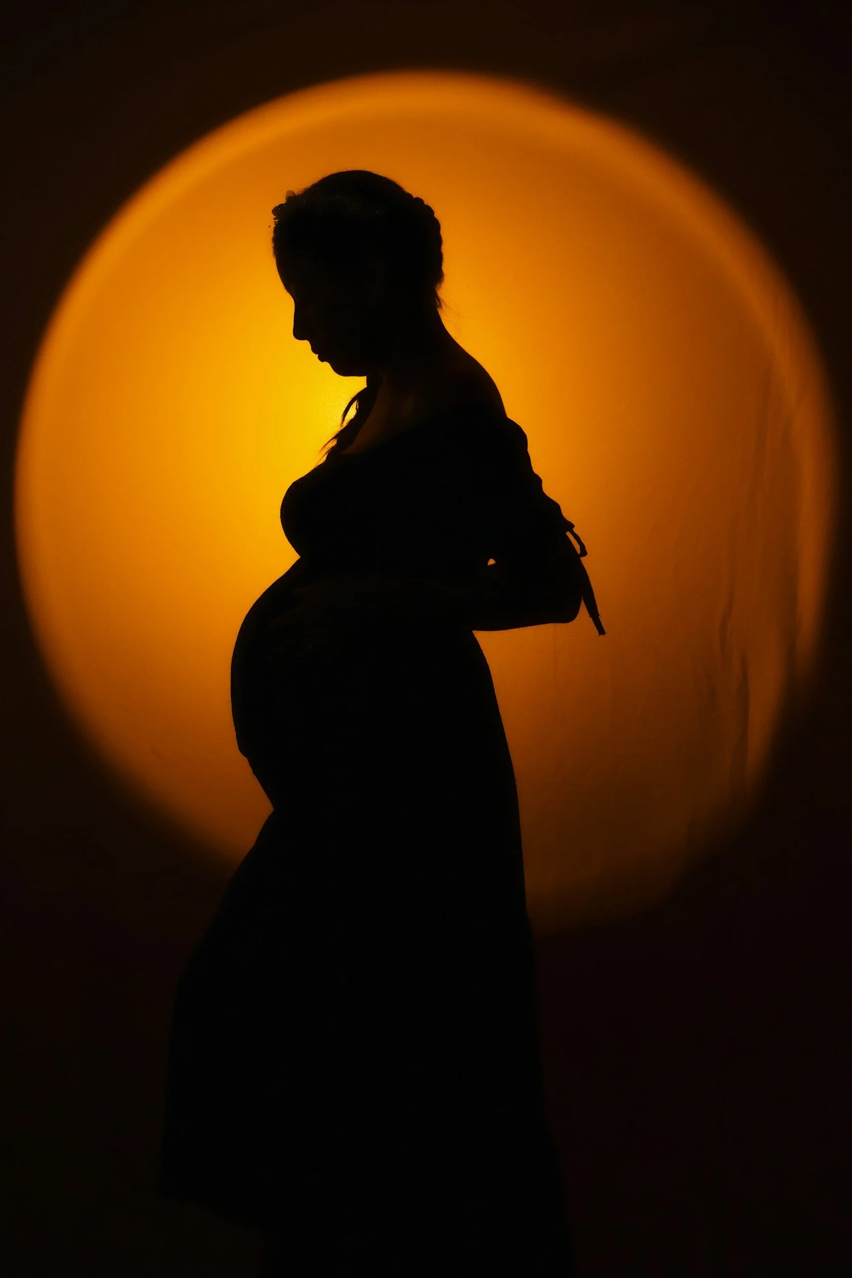 Silhouette of a pregnant woman standing in front of a warm yellow-orange circular backlight.