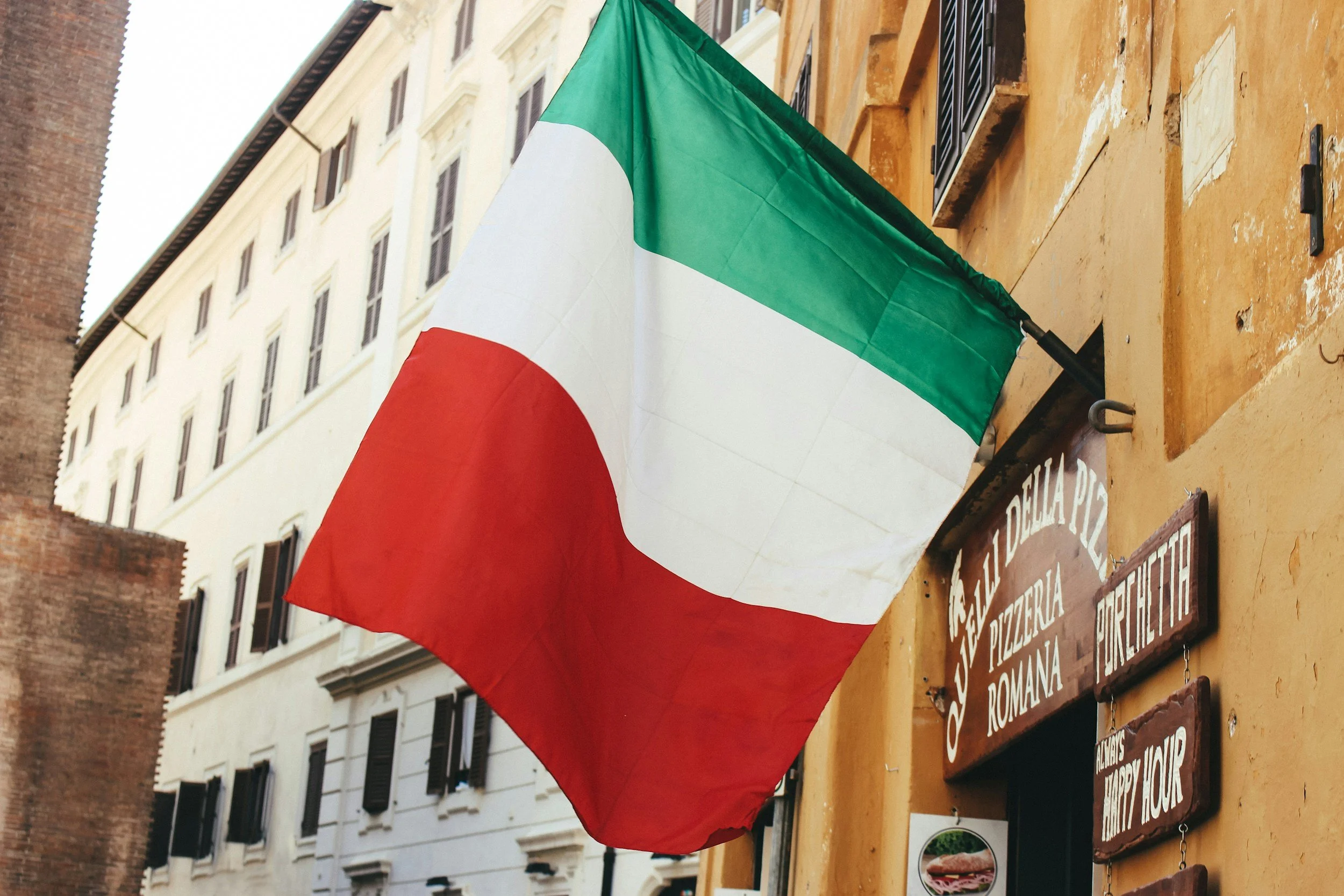 Italian flag hanging outside building with yellow and white walls, black window shutters, and restaurant signage.