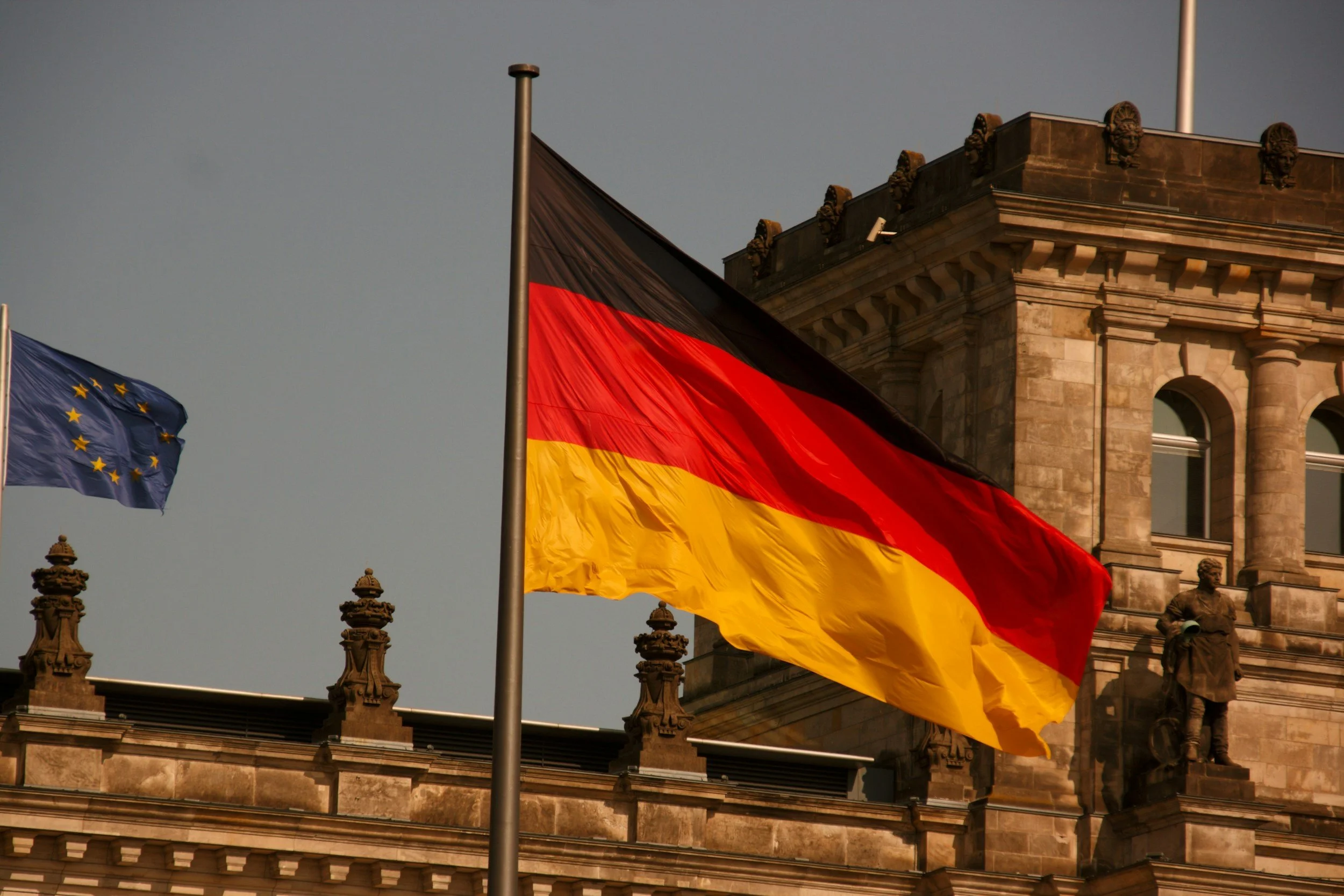 Three flags flying in front of a historic stone building. The flags include the European Union flag, the German flag, and a third flag partially visible.