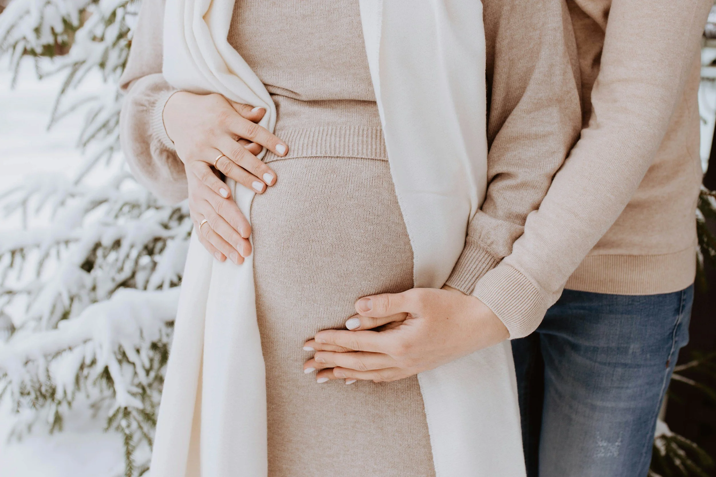 Two women in beige sweaters, with one pregnant, standing outdoors in a snowy setting, holding their bellies with their hands intertwined.