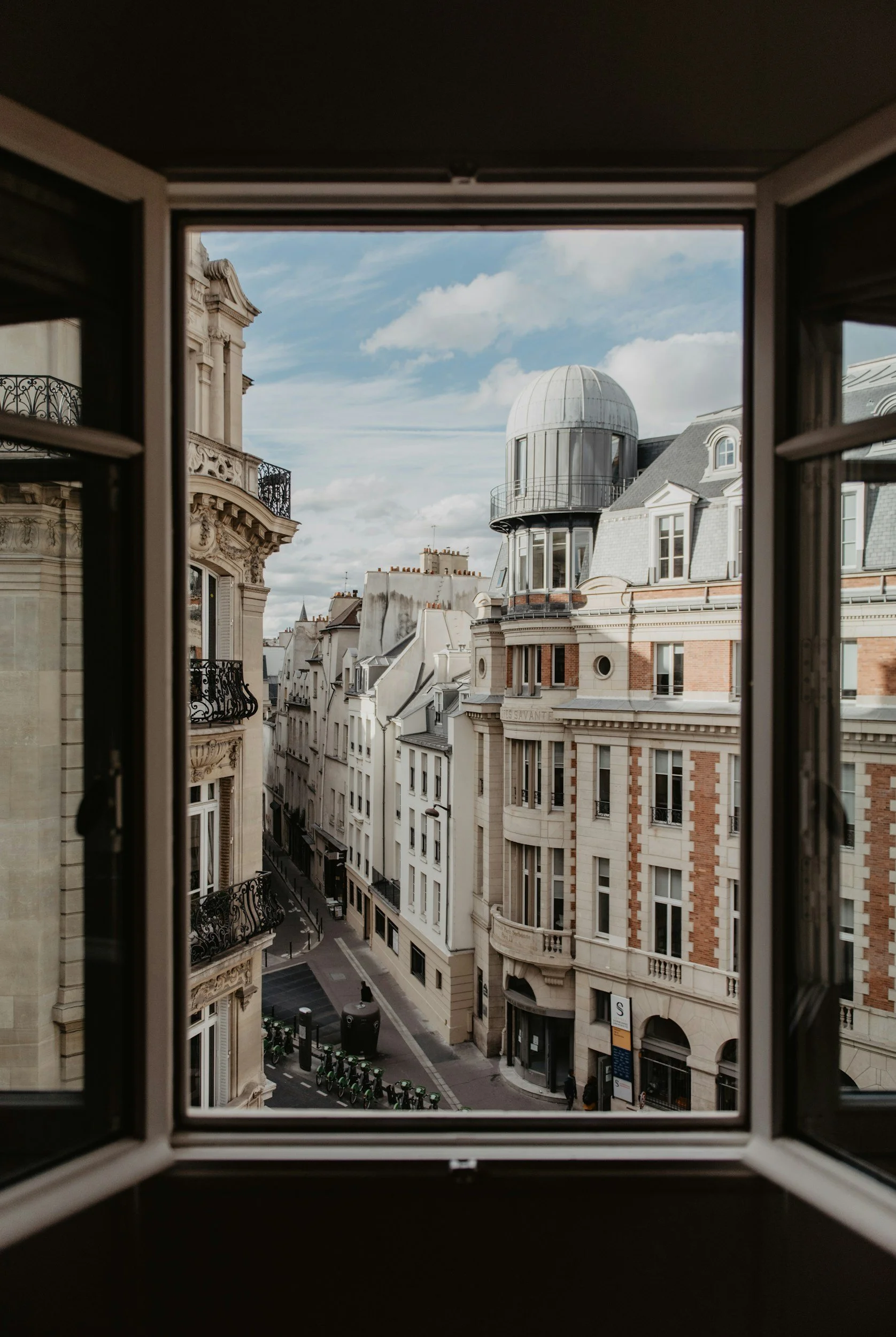 View of European city street from a window, featuring historic buildings with ornate facades, balconies, and a rounded turret, under a partly cloudy sky.