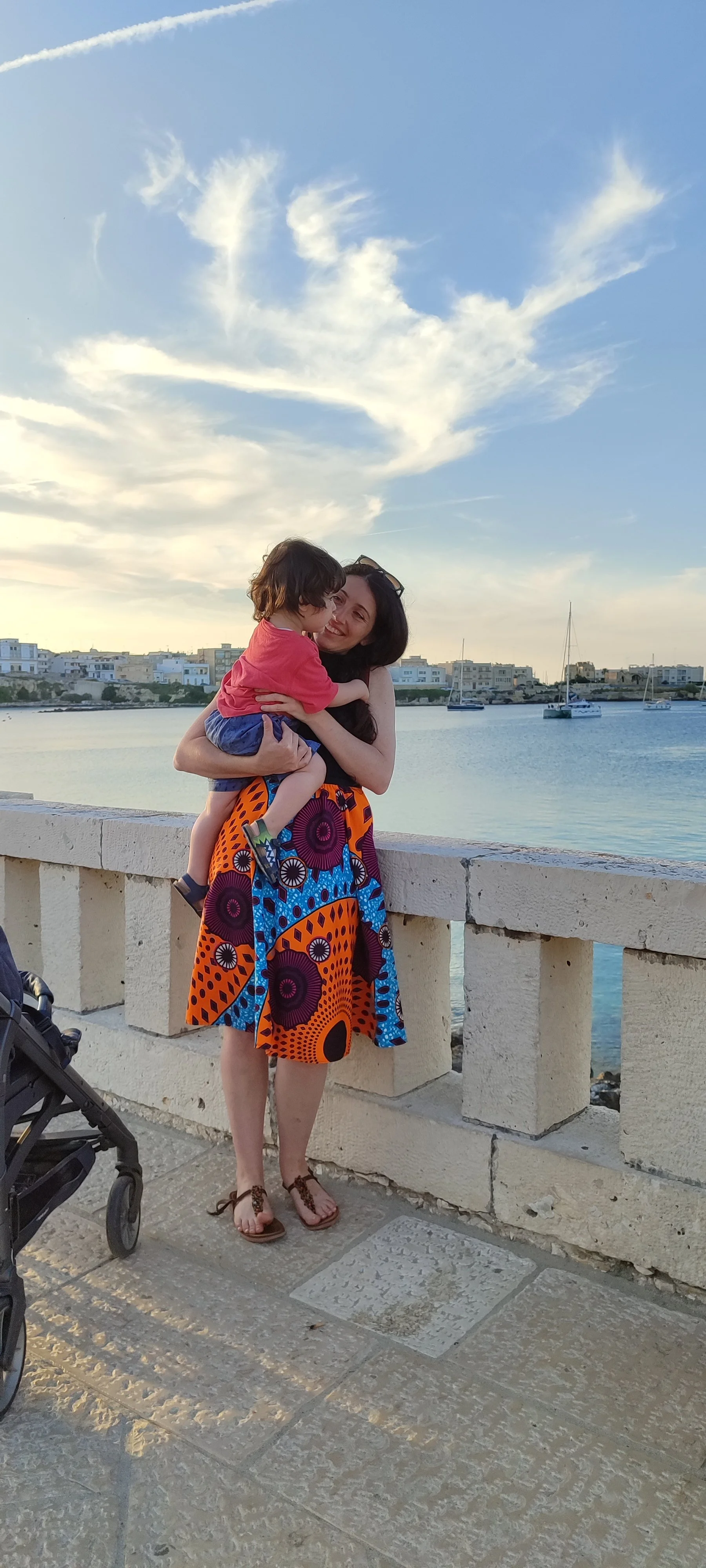 A woman holding a child by a harbor with sailboats, wearing a colorful patterned skirt, sandals, smiling, in front of a concrete railing, with a stroller partially visible on the left, under a blue sky with wispy clouds.