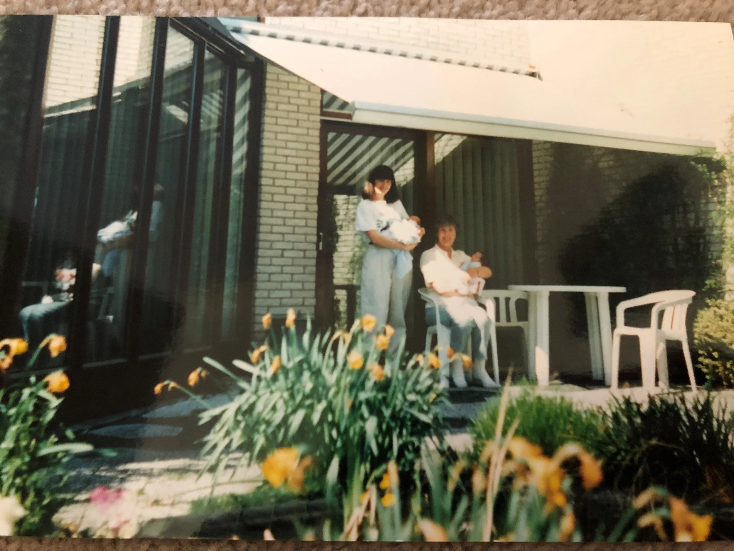 A woman holding a newborn baby and sitting on a chair on a porch, with another woman standing beside her holding a baby, on a sunny day. There are flowers in the foreground and a patio area behind them.