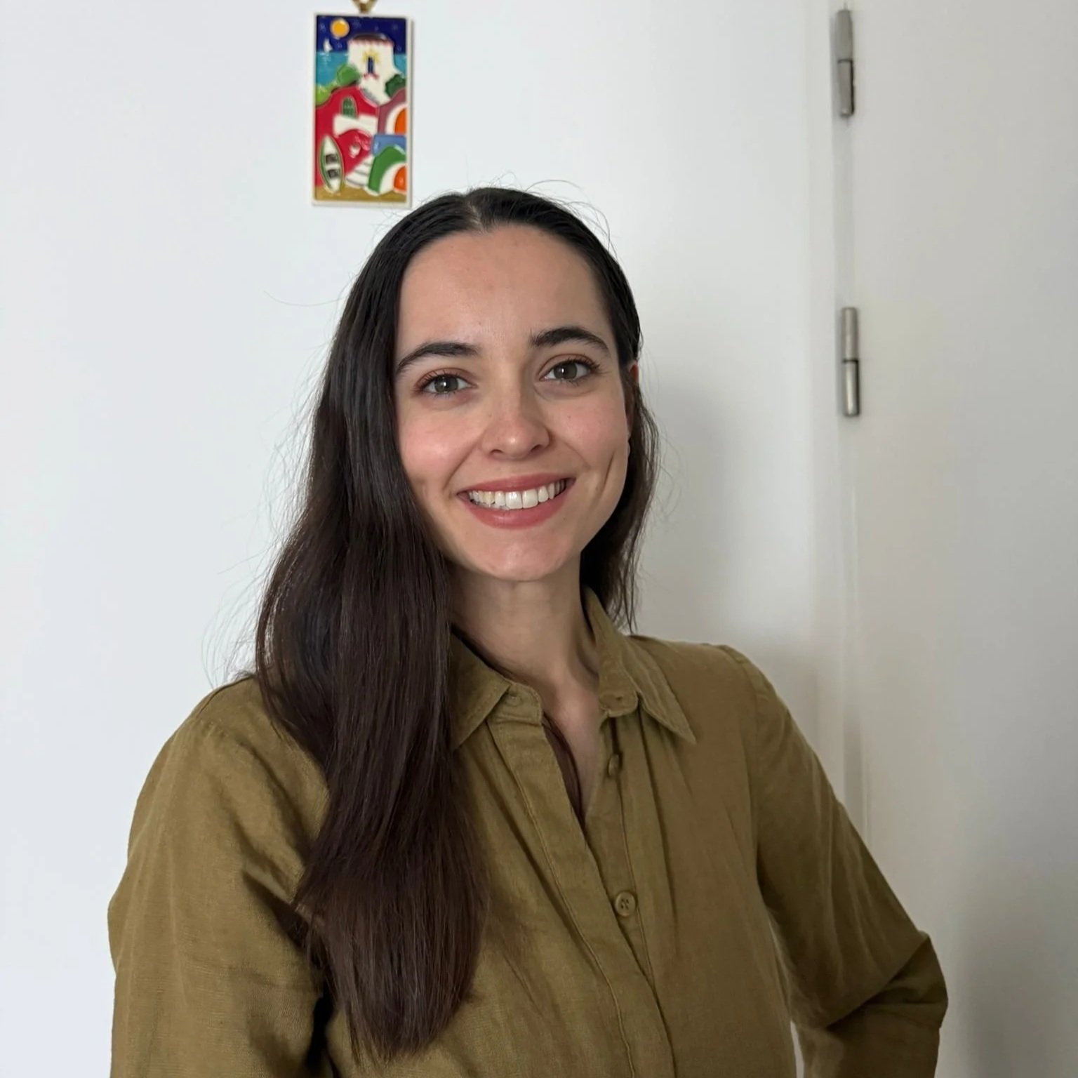 A young woman with long dark hair, smiling, wearing a khaki button-up shirt, standing in front of a white wall with a colorful abstract painting hanging behind her.