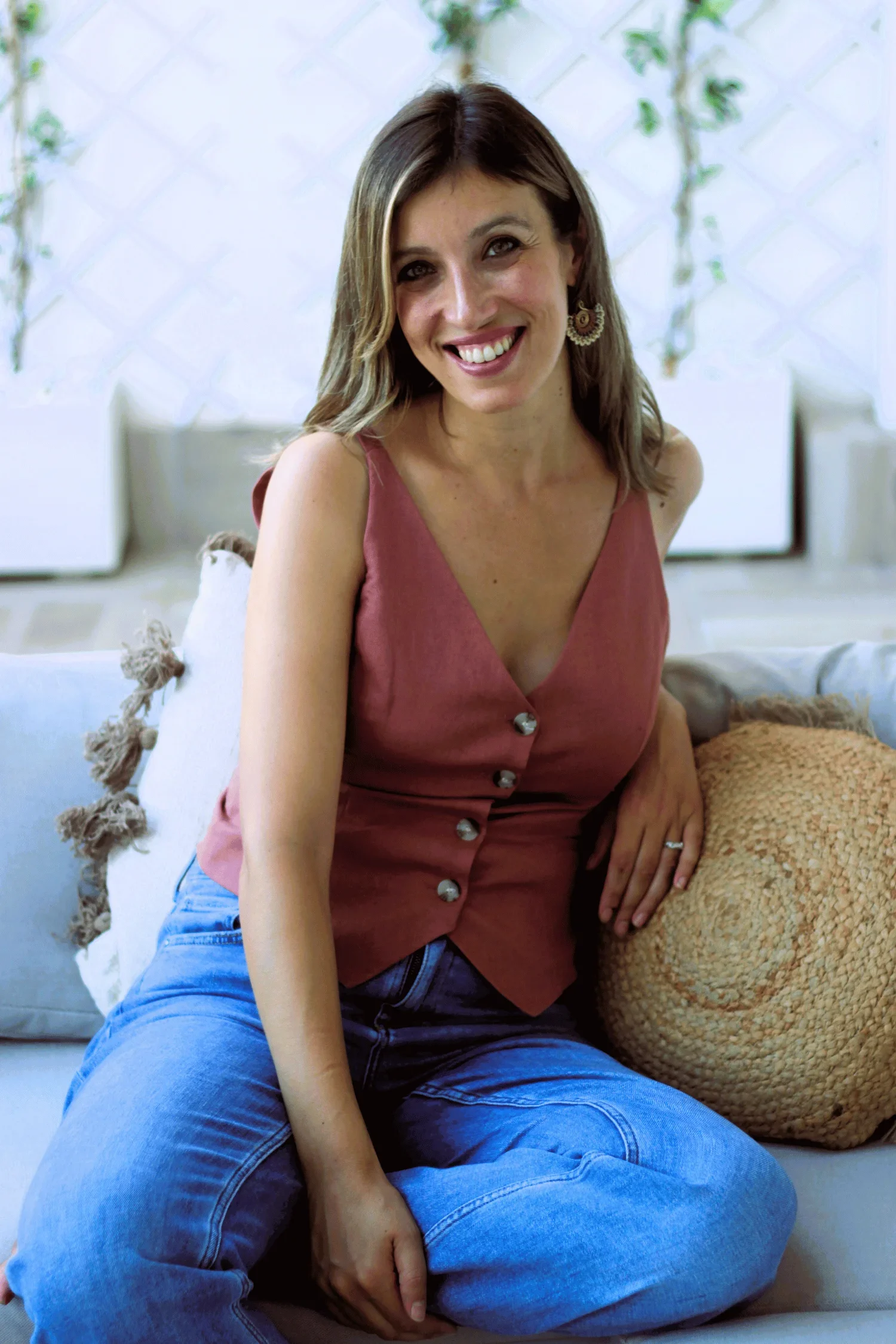 A woman with shoulder-length brown hair, wearing a sleeveless pink top with buttons, smiling and sitting on a sofa with a decorative pillow beside her. The background features a white wall with a lattice pattern and hanging green plants.