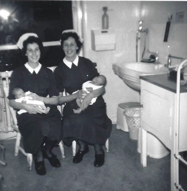 Two women, dressed in dark uniforms with white collars, sitting in a hospital room holding two newborn babies.