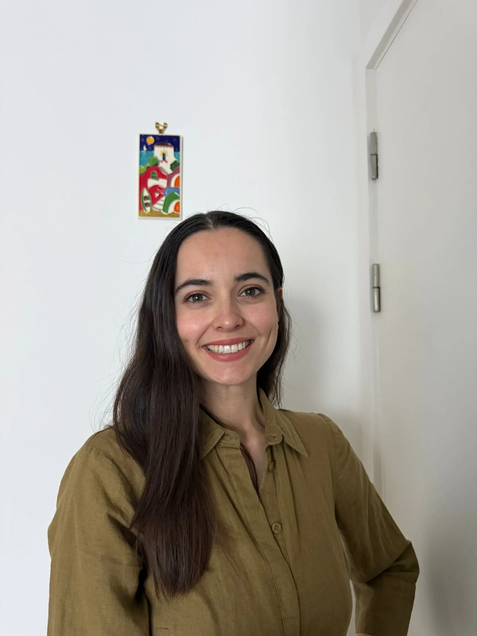 A woman with long dark hair smiling and wearing a brown button-up shirt, standing in front of a white wall with a colorful abstract artwork hanging behind her.