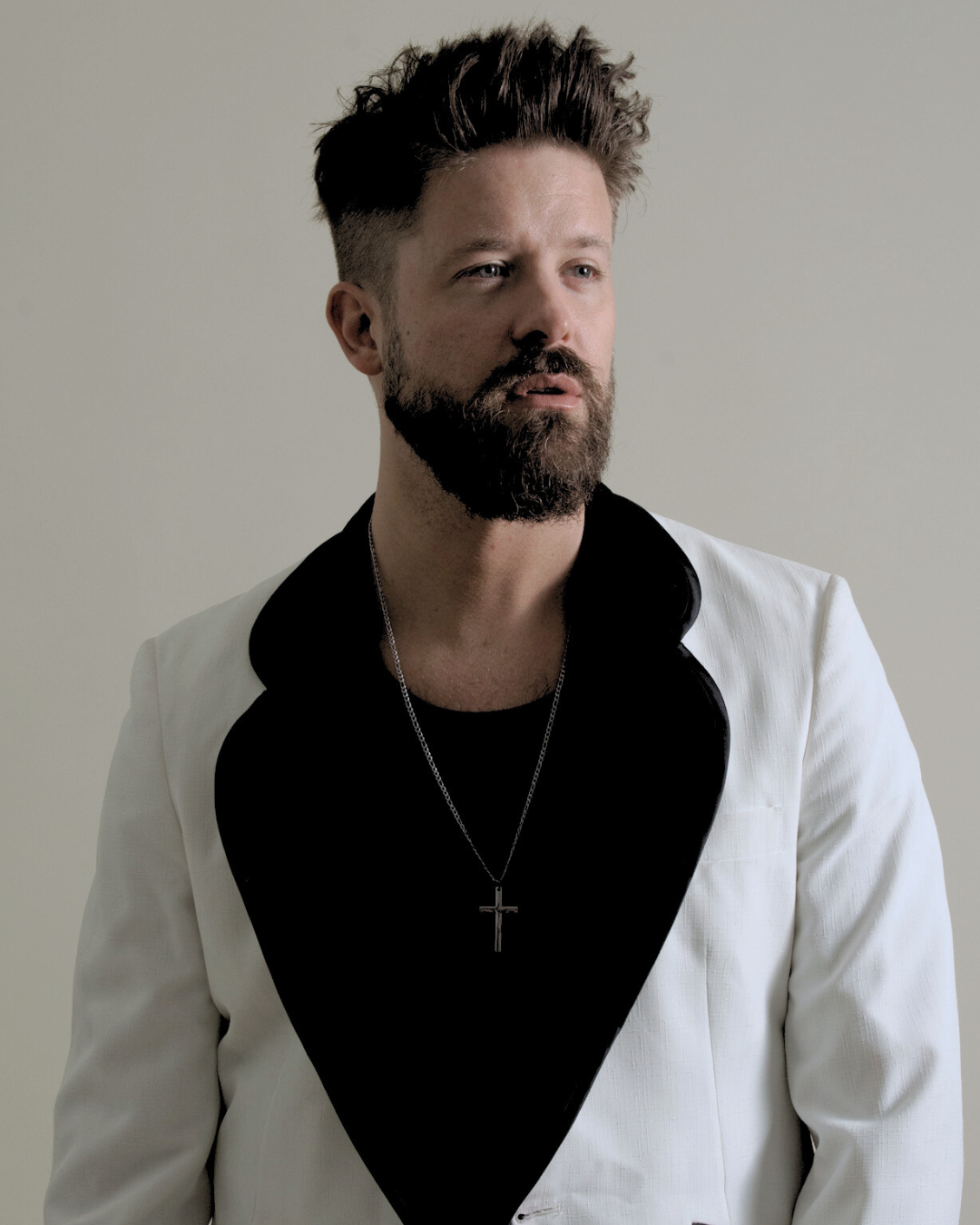 A man with dark, styled hair and a beard, wearing a white blazer with black accents, a black shirt, and a silver cross necklace, looking to the side against a plain, light-colored background.