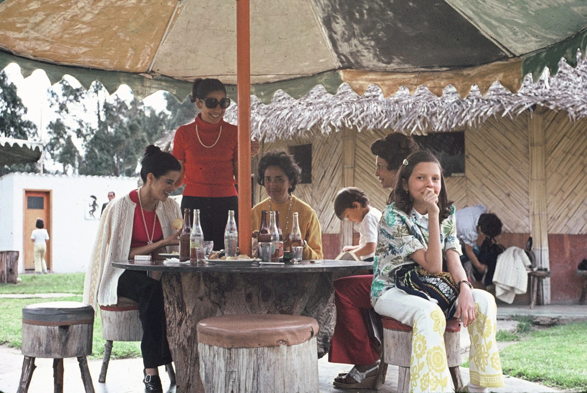A group of women and children enjoying a meal outdoors under a thatched roof. The women are dressed in colorful clothing and accessories, and some are seated at a round wooden table with drinks and food. A woman in red with sunglasses is standing and smiling, while another woman in a floral dress is sitting and looking at the camera.