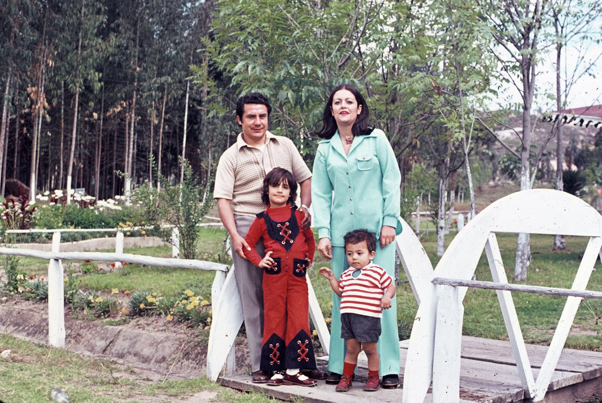 A woman with dark hair and a man with dark hair stand on a small white bridge with three children in a garden or park with trees and flowers, during daytime.