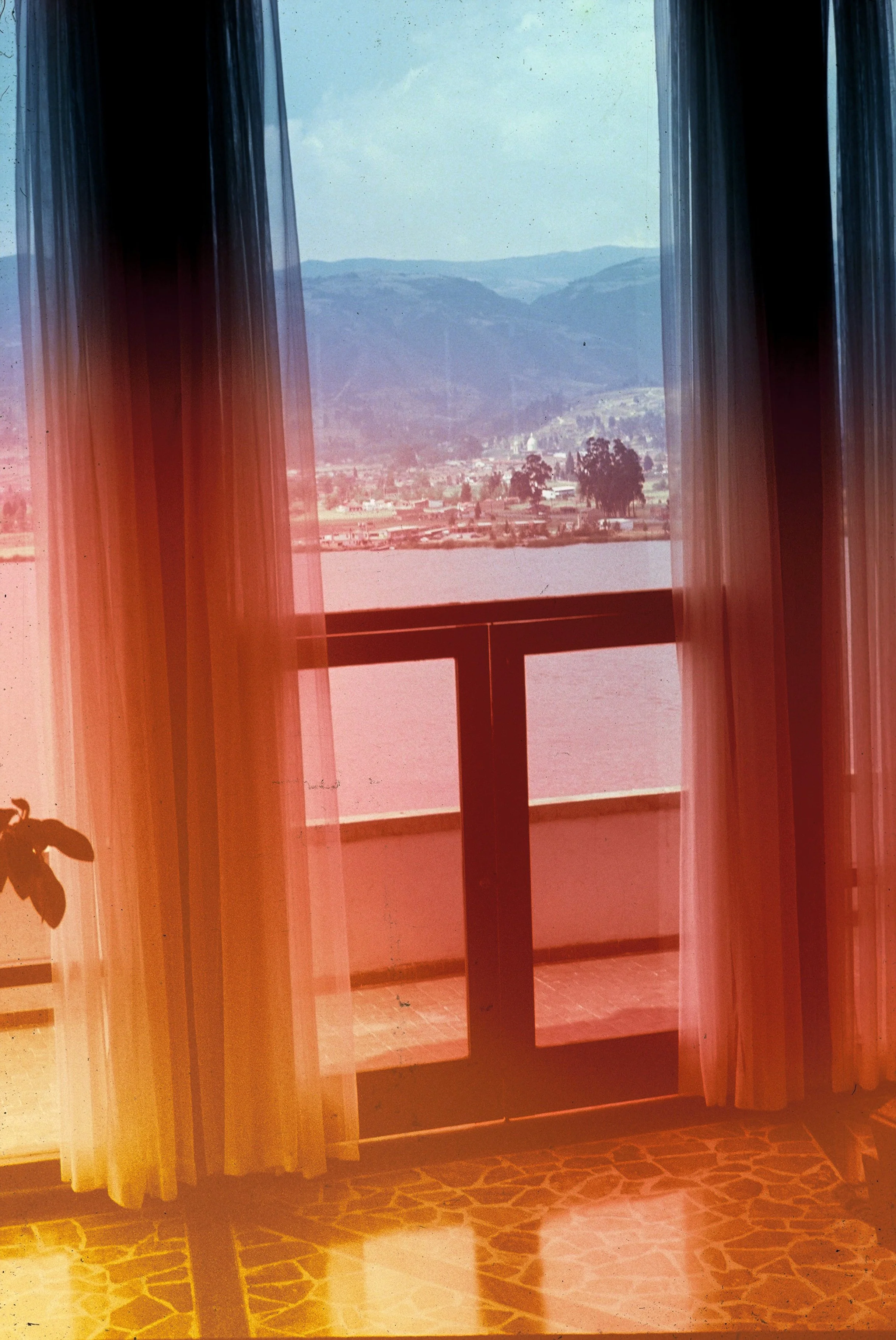 View of mountains and a town seen through a window with pinkish curtains, taken with warm, reddish lighting.