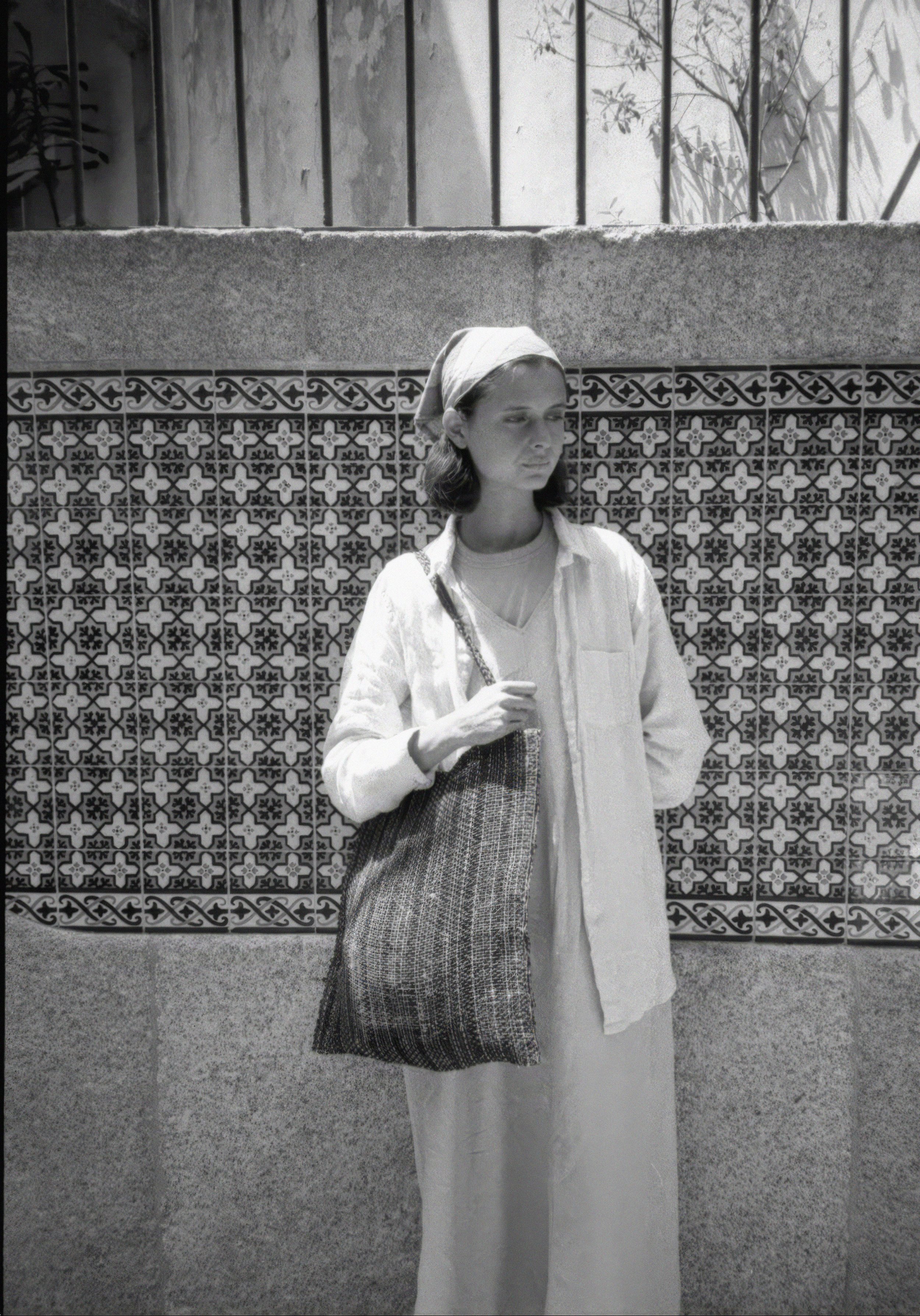 A woman wearing a headscarf, white shirt, and long skirt standing against a decorative tiled wall. She holds a bag in her left hand and looks down. The background shows a wall with patterned tiles and a concrete section with some plants and a metal fence above.