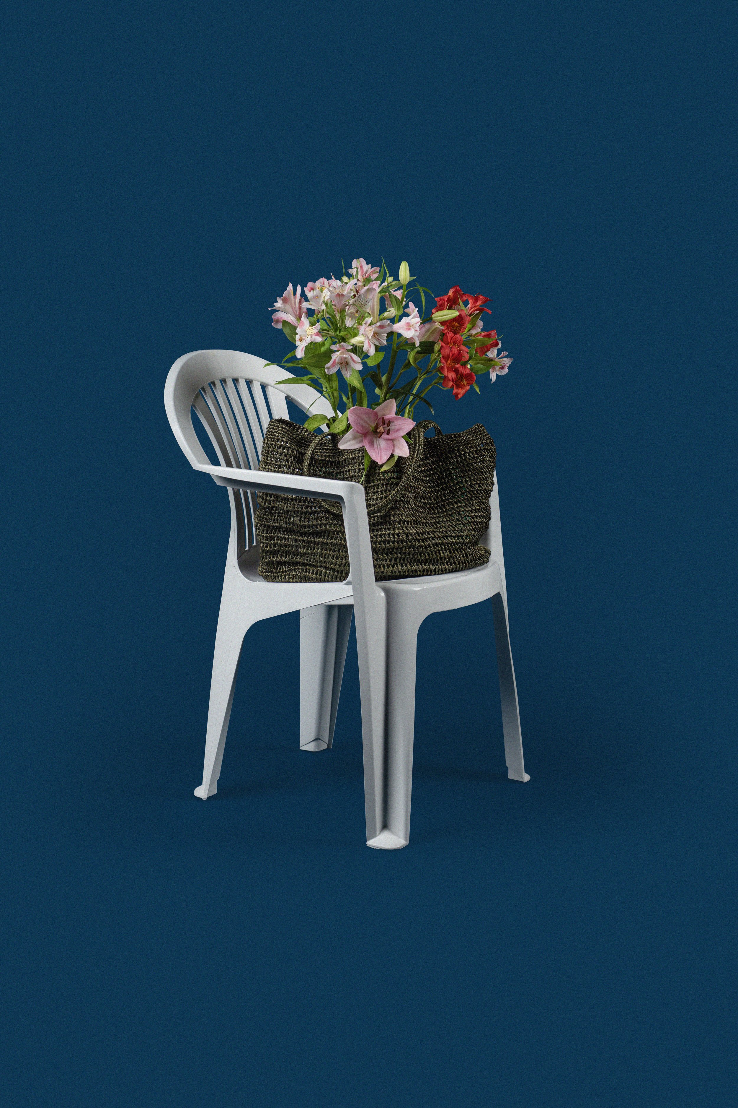 A white plastic chair with a woven bag on top, holding a bouquet of pink and red flowers, set against a plain blue background.