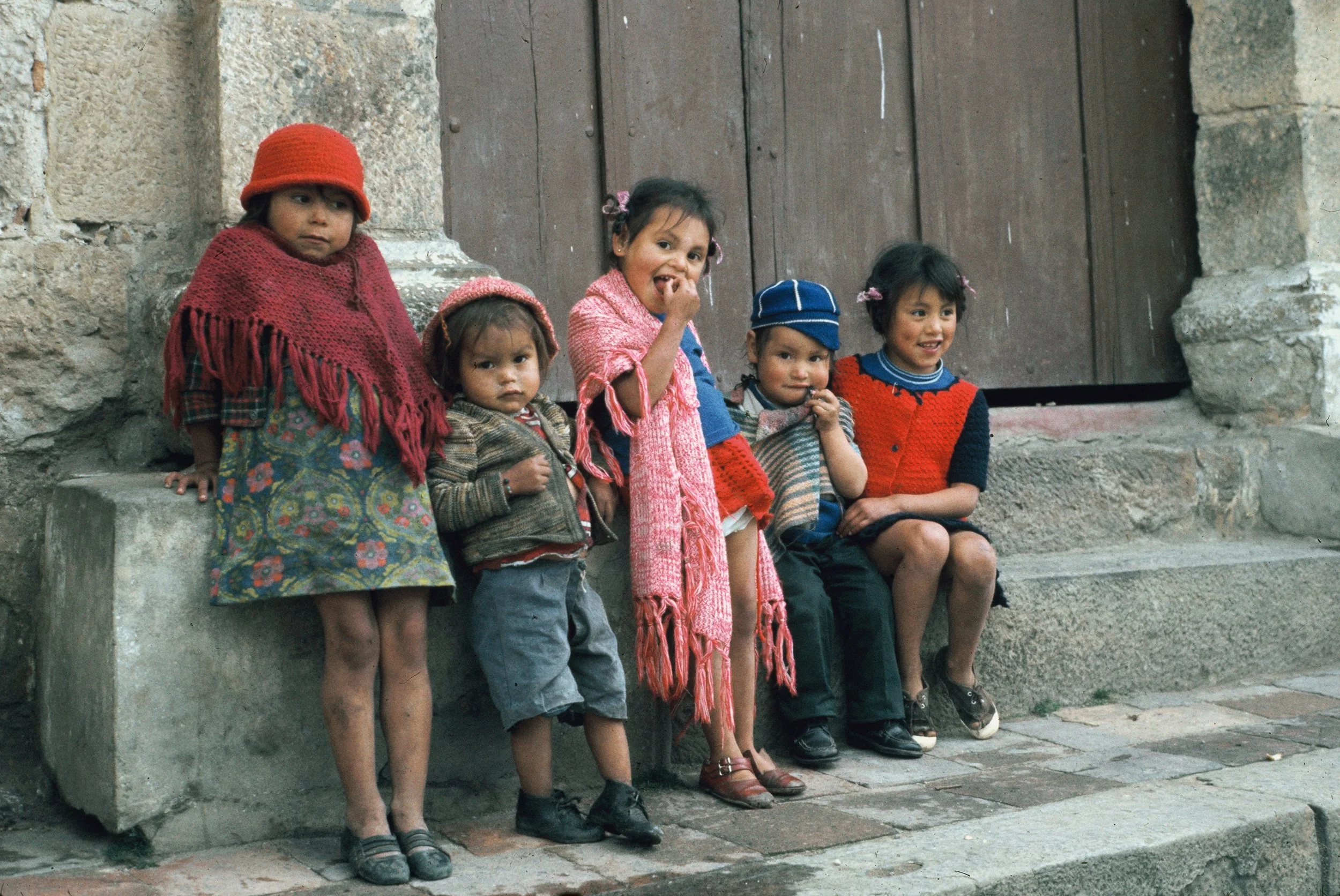 A group of six children standing and sitting on a step outside a stone building with a wooden door, dressed in colorful clothing and looking at the camera.