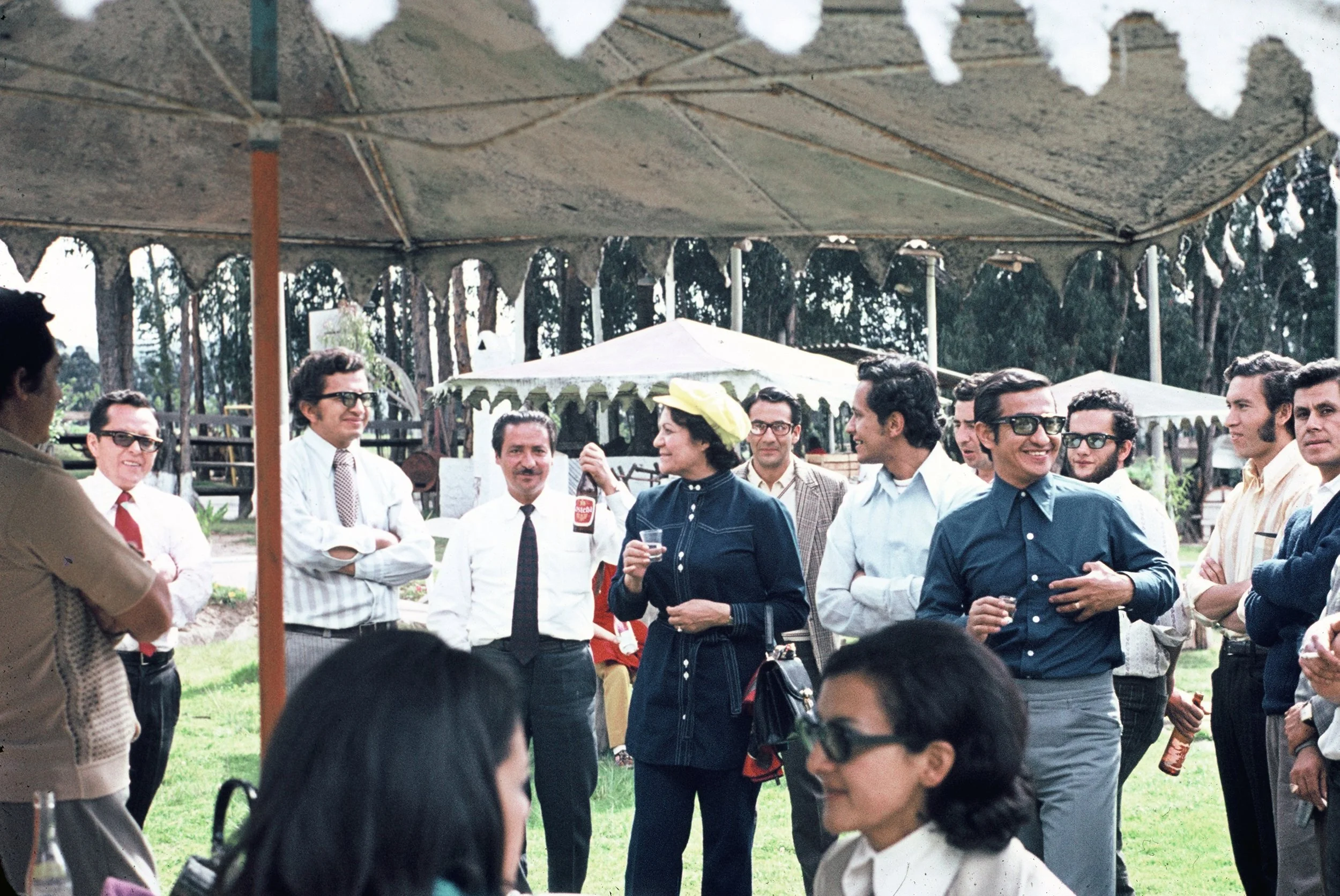 A group of people, mostly men, are gathered outdoors in a social setting under a canopy, wearing vintage clothing and glasses, holding drinks and engaging in conversation at a party or celebration.