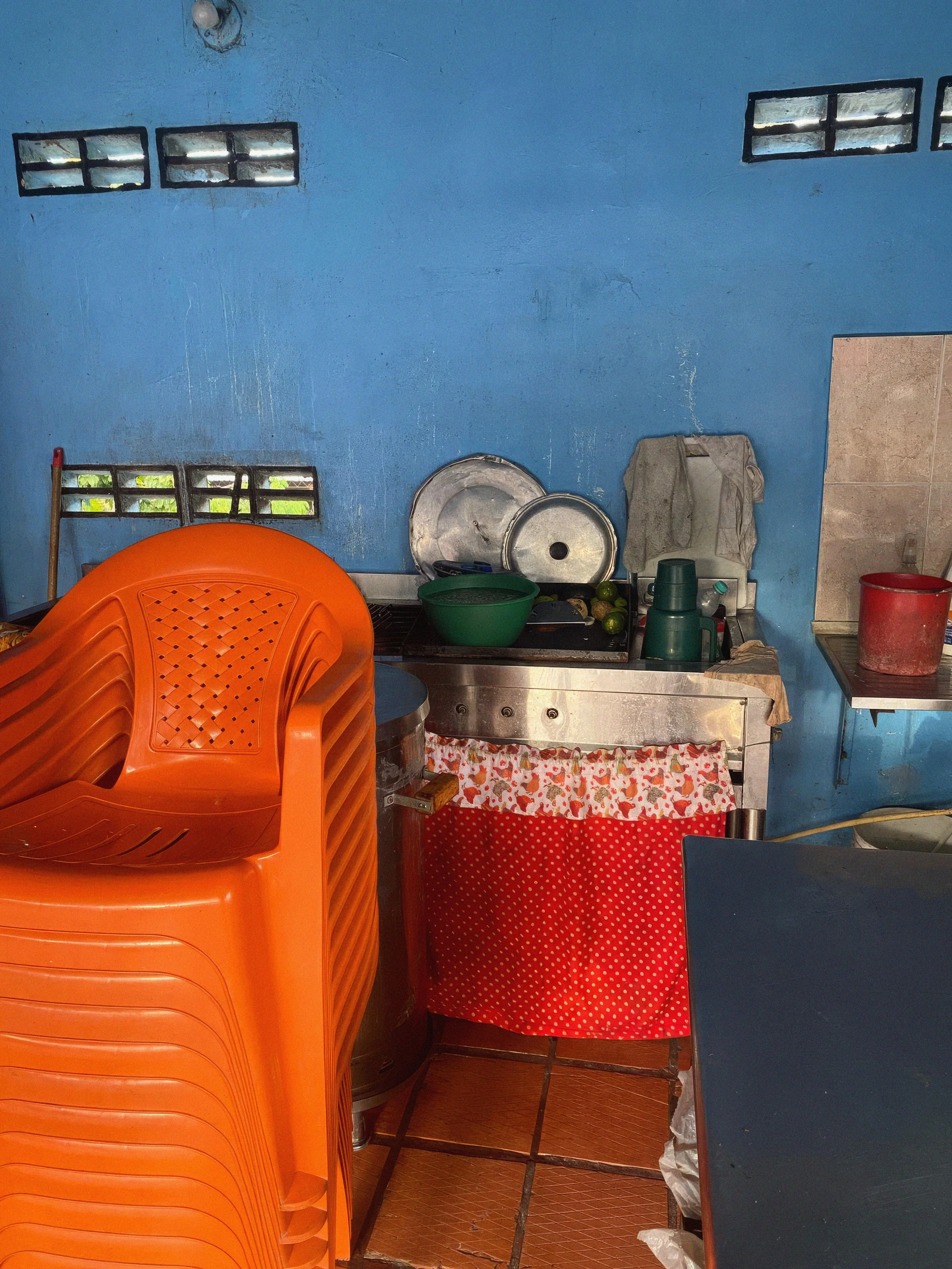 A small kitchen with a light blue wall, small rectangular windows, orange plastic chairs stacked, a green bowl, metal pots, and a red and white polka dot curtain covering some storage below the counter.