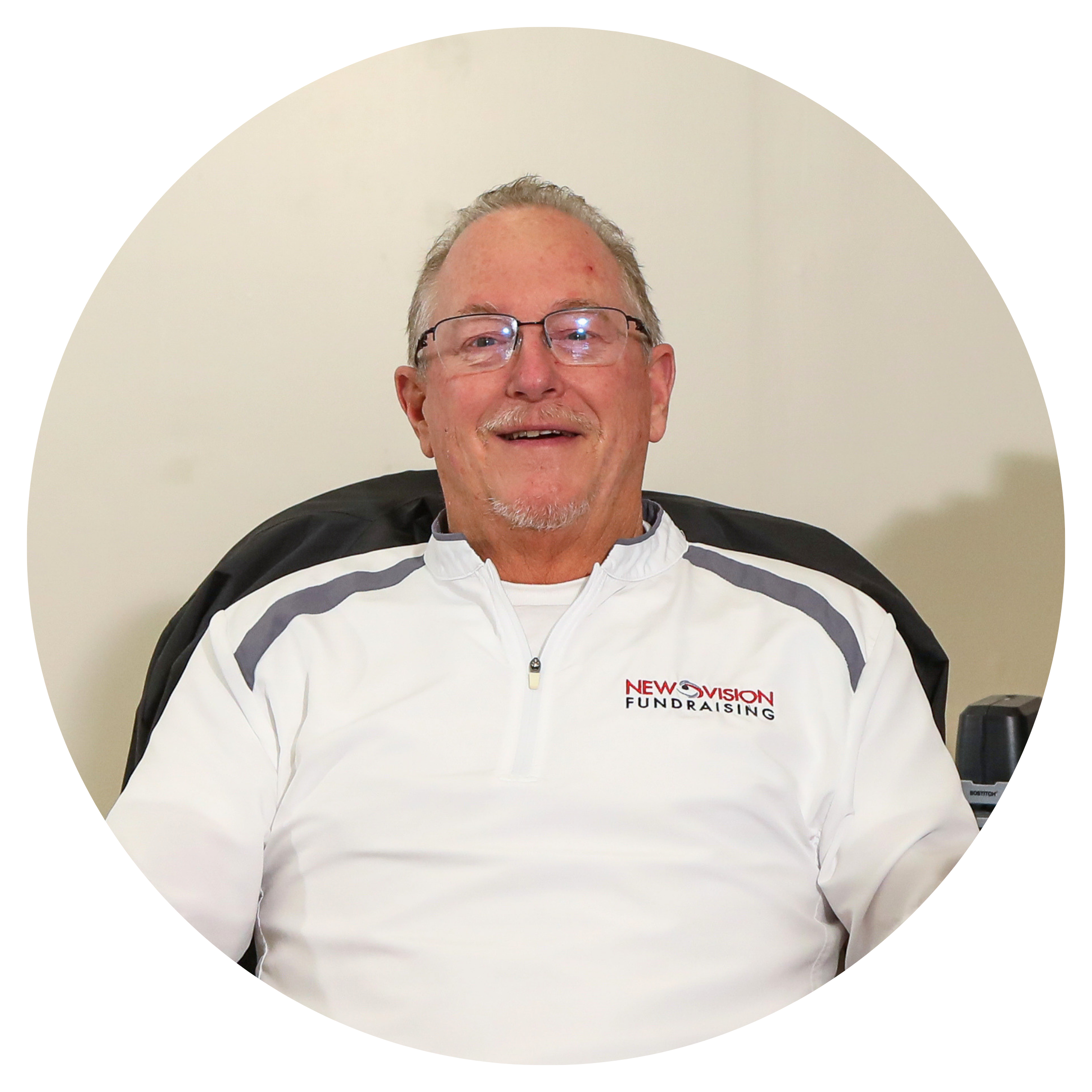 A man with glasses smiling, wearing a white jacket with the text 'NEW VISION FUNDRAISING' on it, sitting in front of a plain, light-colored wall.