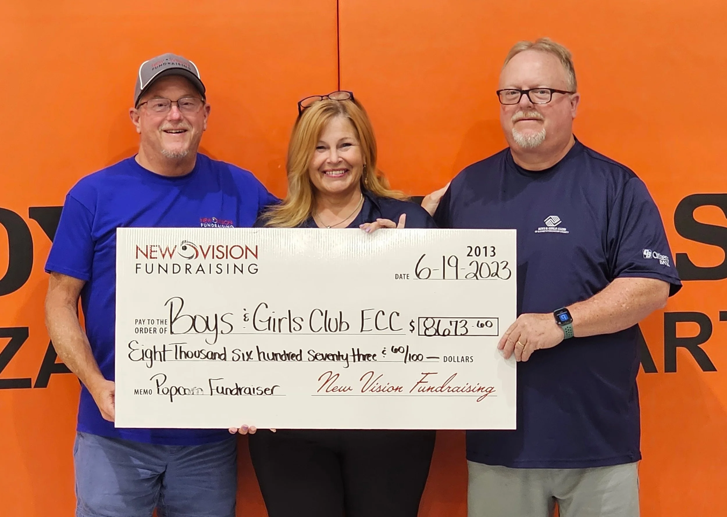 Three people holding a large check at an event with an orange background. The check is made out to Boys and Girls Club ECC for $8,673.60, dated June 19, 2023.